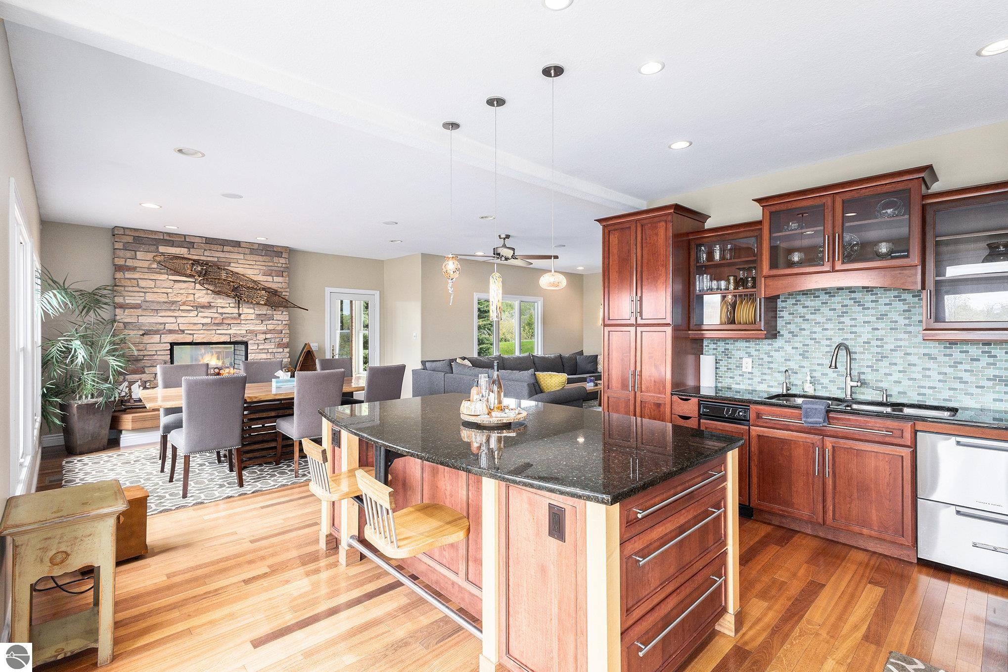 Open-plan kitchen with a granite island, wood cabinetry, and stone fireplace beside the dining and living areas.