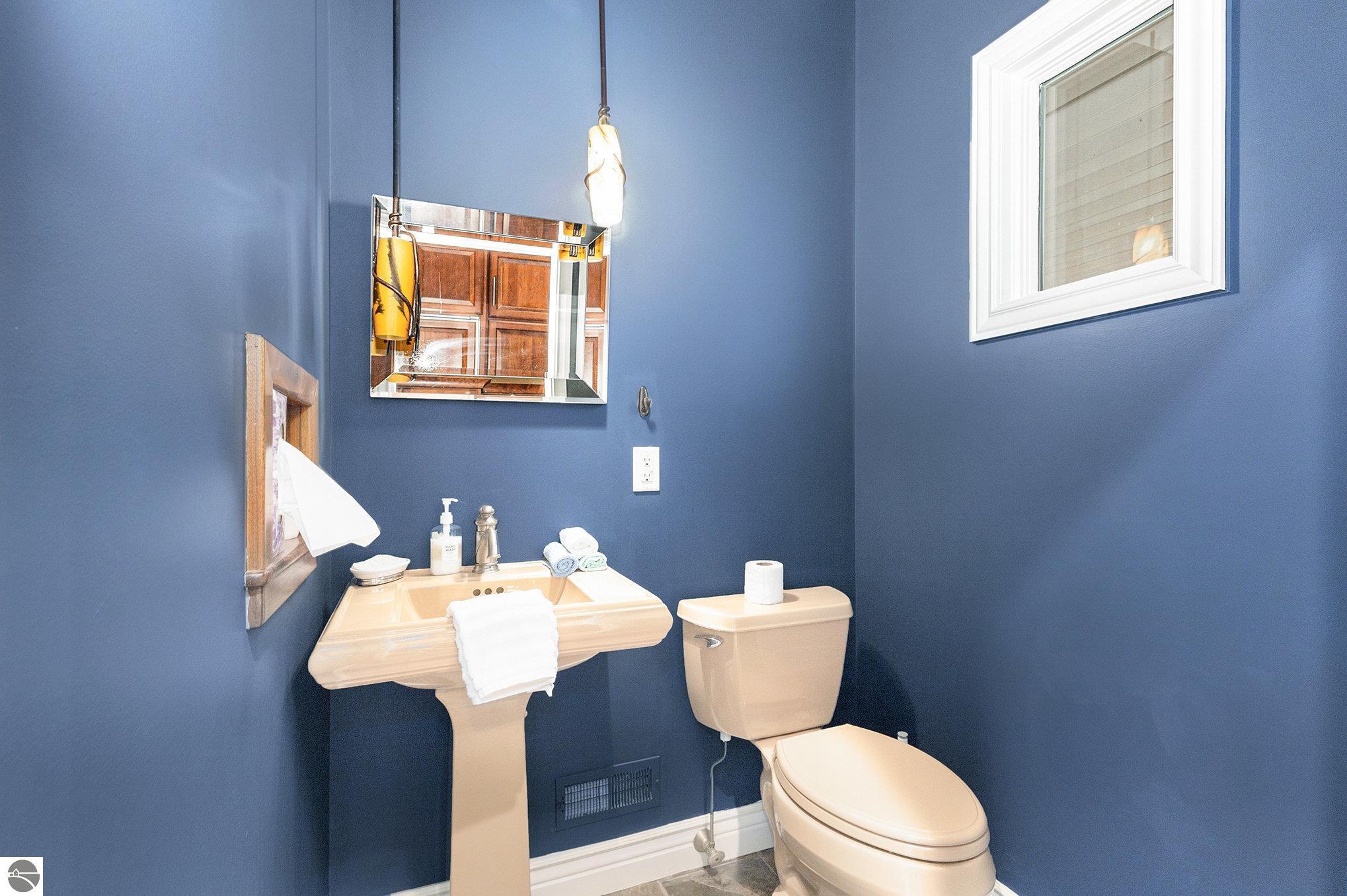 Bathroom with deep blue walls, beige pedestal sink and toilet, framed mirror, and high window.
