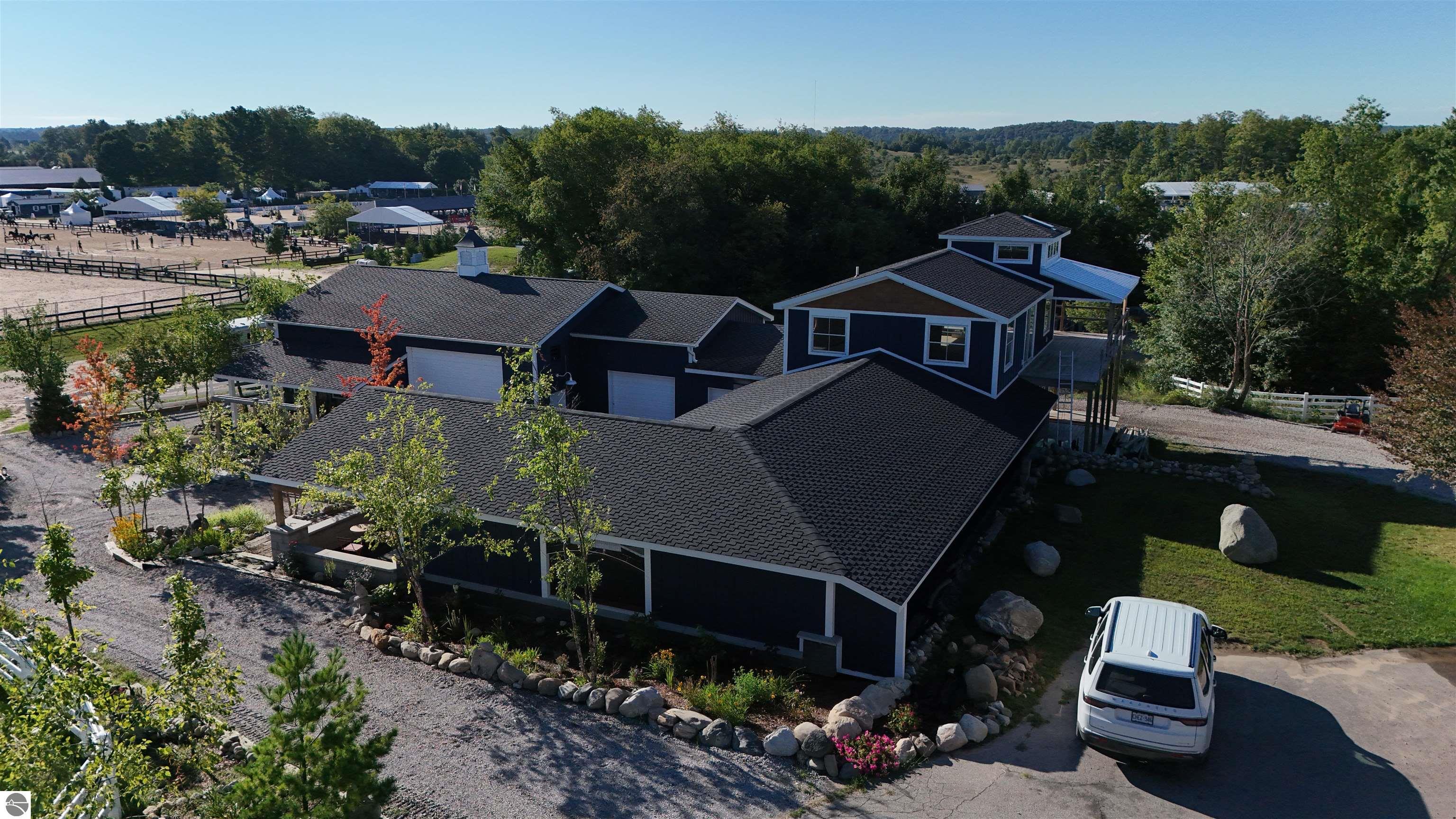 Aerial view of a dark blue equestrian property beside fenced riding arenas and wooded hills.