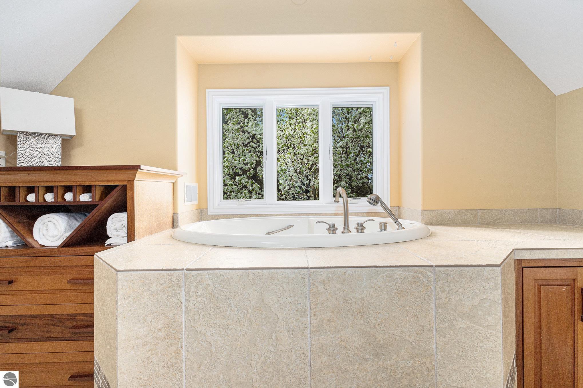 Oval soaking tub in a raised tiled platform beneath a window overlooking flowering trees.