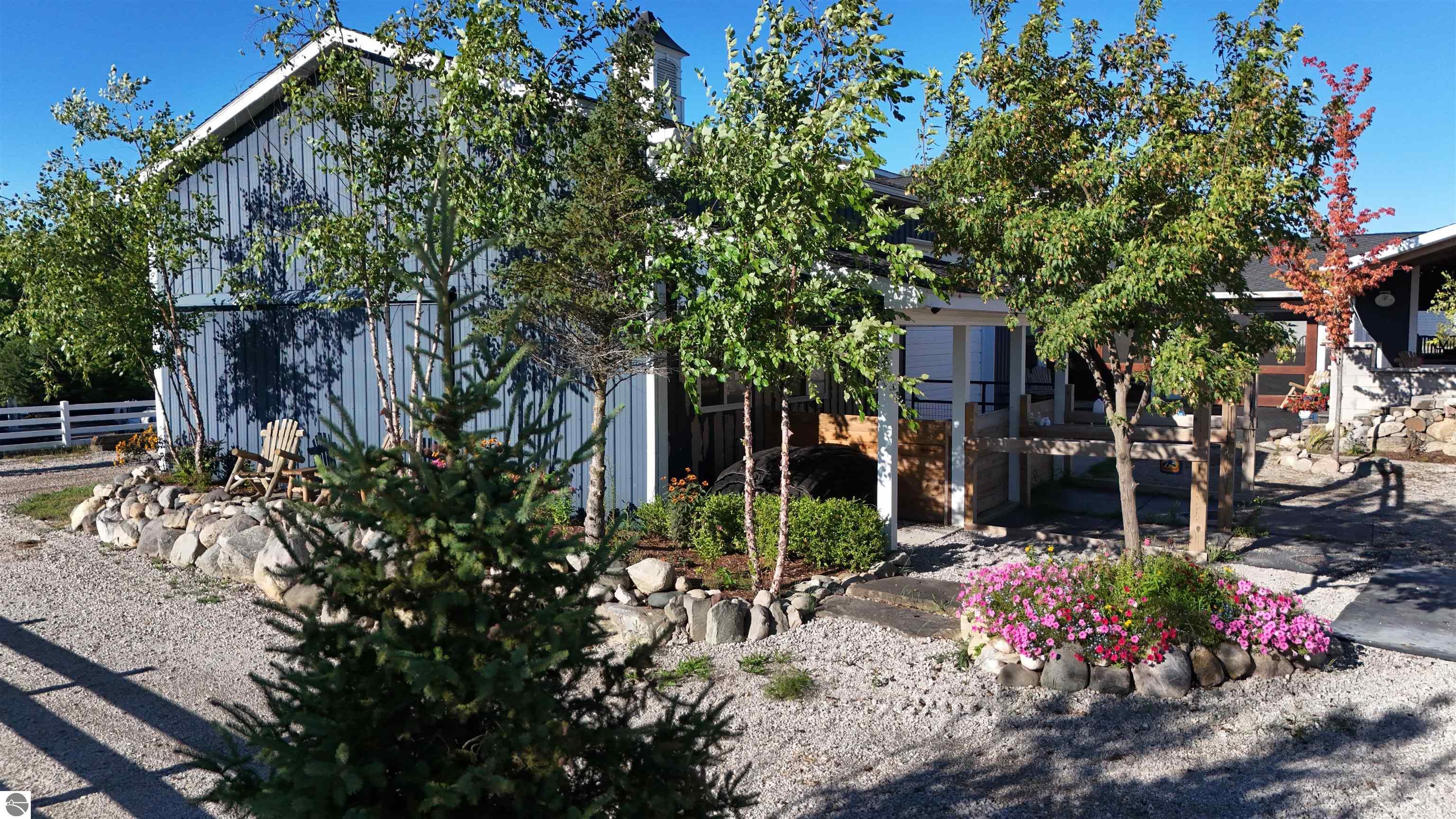 Gray equestrian barn and stable with a covered entry, gravel paths, and pink flower beds.