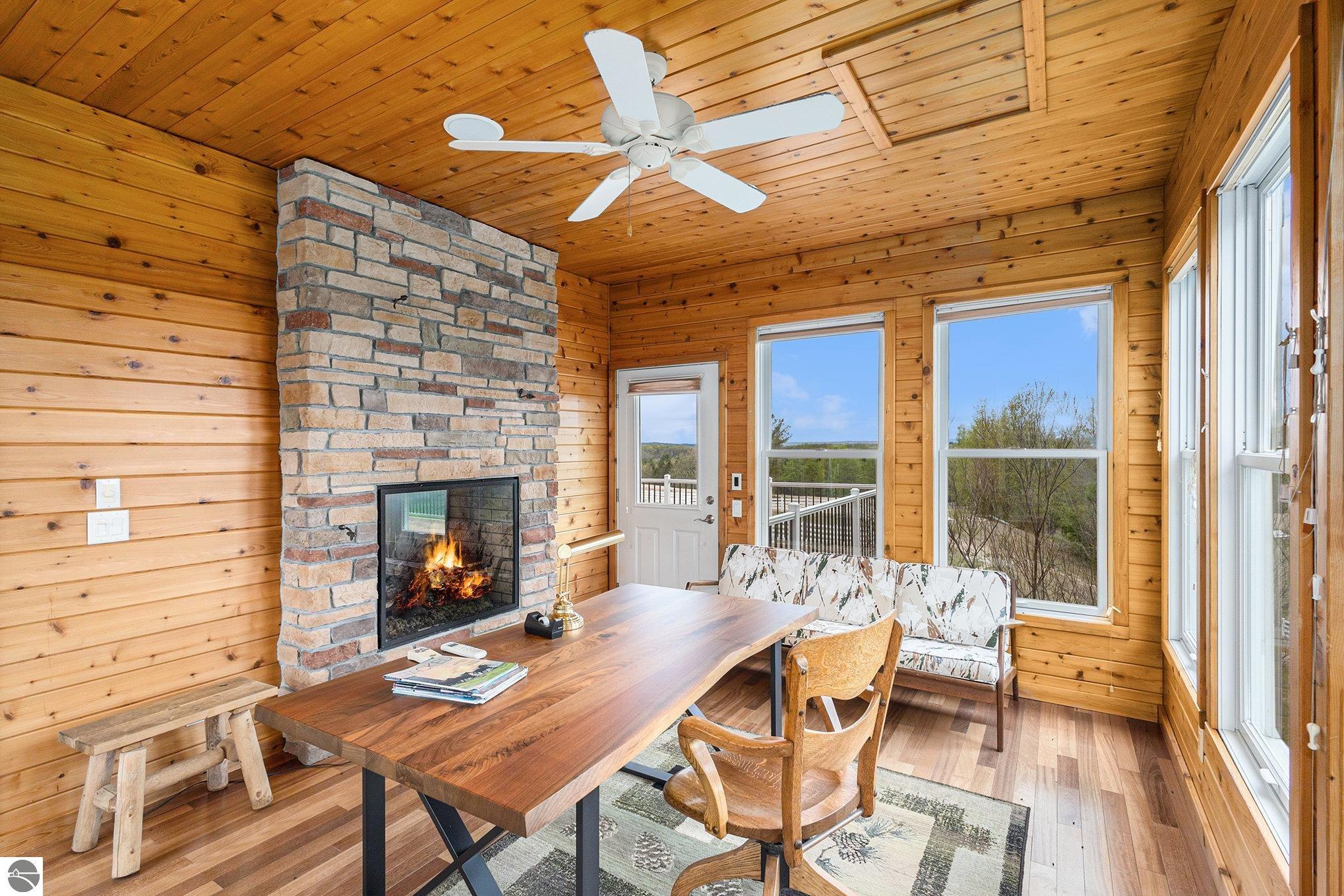 Wood-paneled dining area with a stone fireplace, long table, and large windows overlooking trees.