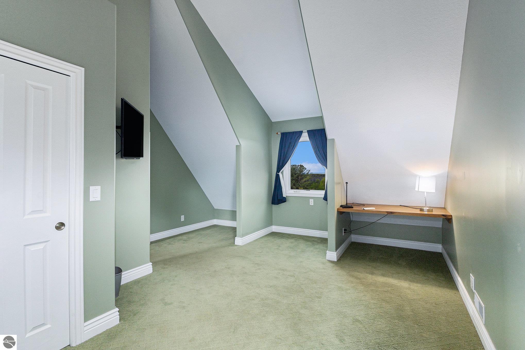 Attic room with a desk, wall-mounted television, and curtained window overlooking trees.