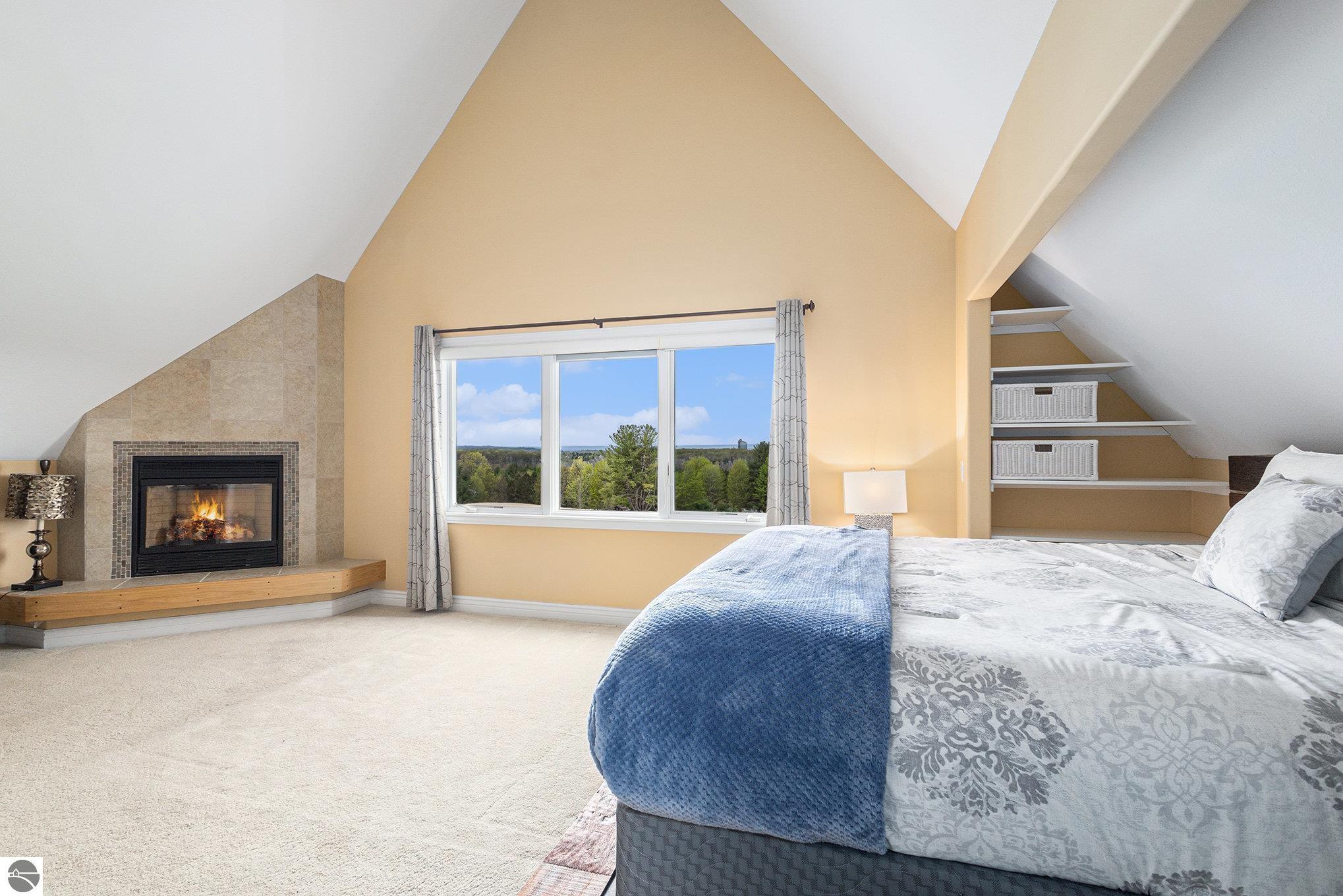Bedroom with vaulted ceiling, tiled fireplace, and a large window overlooking trees.