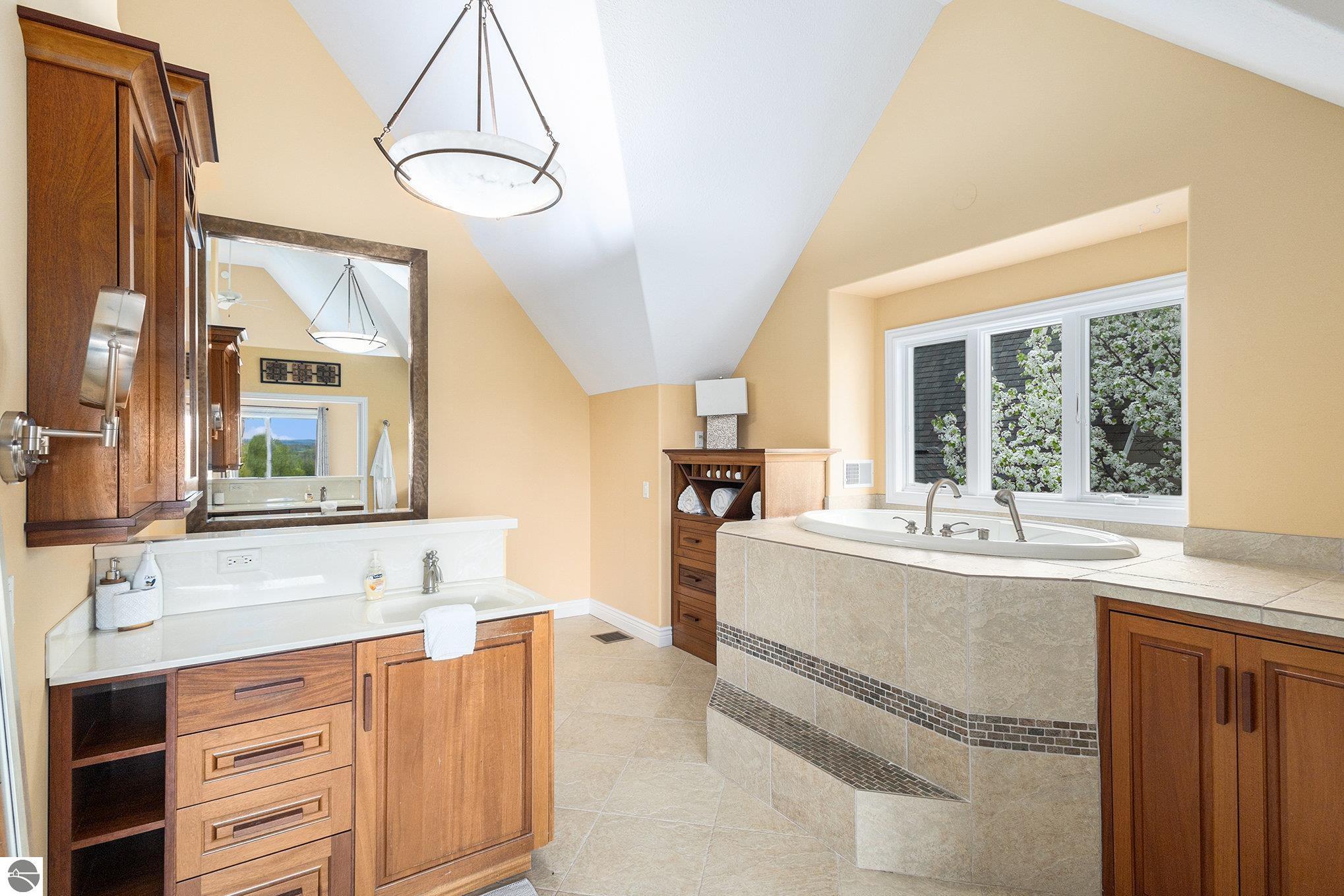 Spacious bathroom with a wood double vanity and tiled soaking tub beside a window of flowering trees.