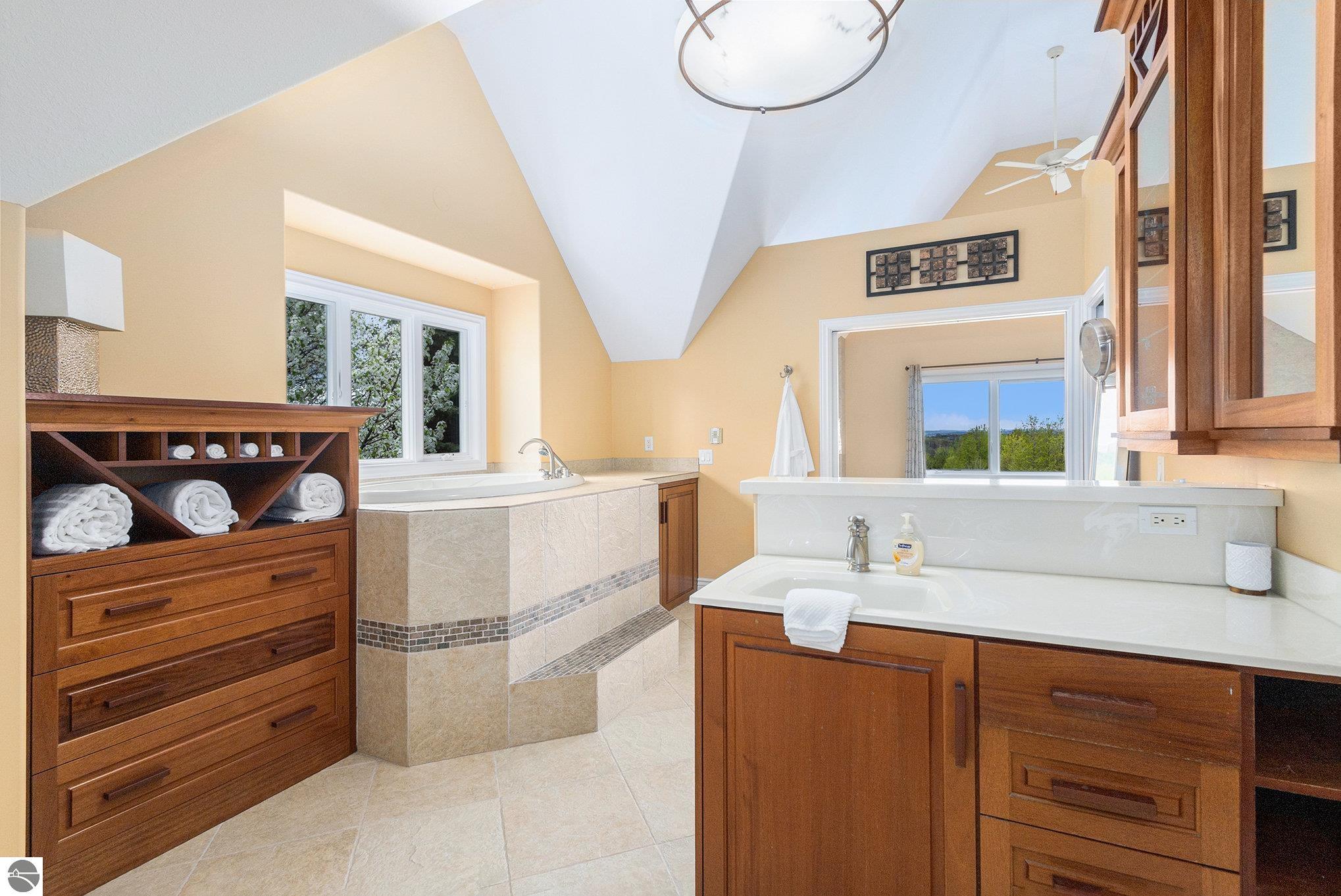 Bathroom with a tiled soaking tub, wood cabinetry, vaulted ceiling, and windows overlooking trees.