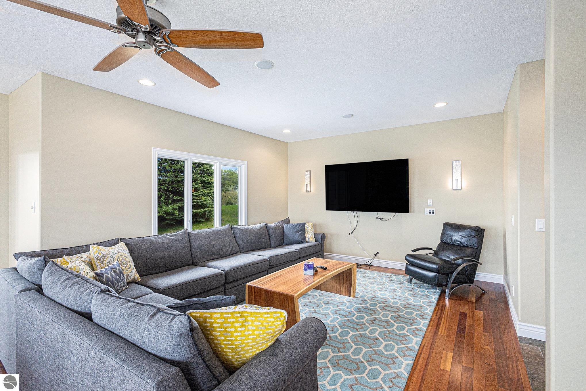Living room with a gray sectional, wall-mounted television, and windows overlooking greenery.