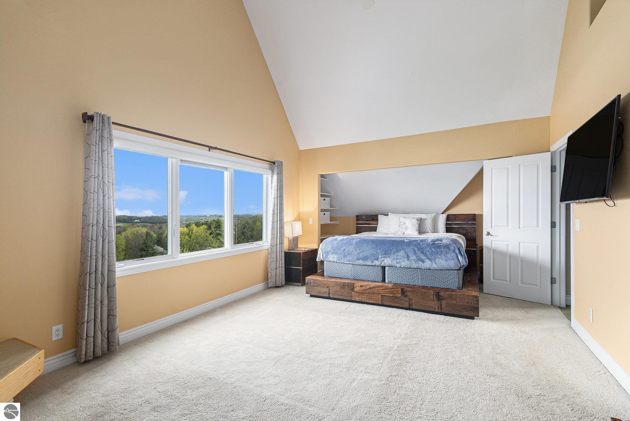 Furnished bedroom with a vaulted ceiling, wooded views through large windows, and a bed on a rustic wood platform.