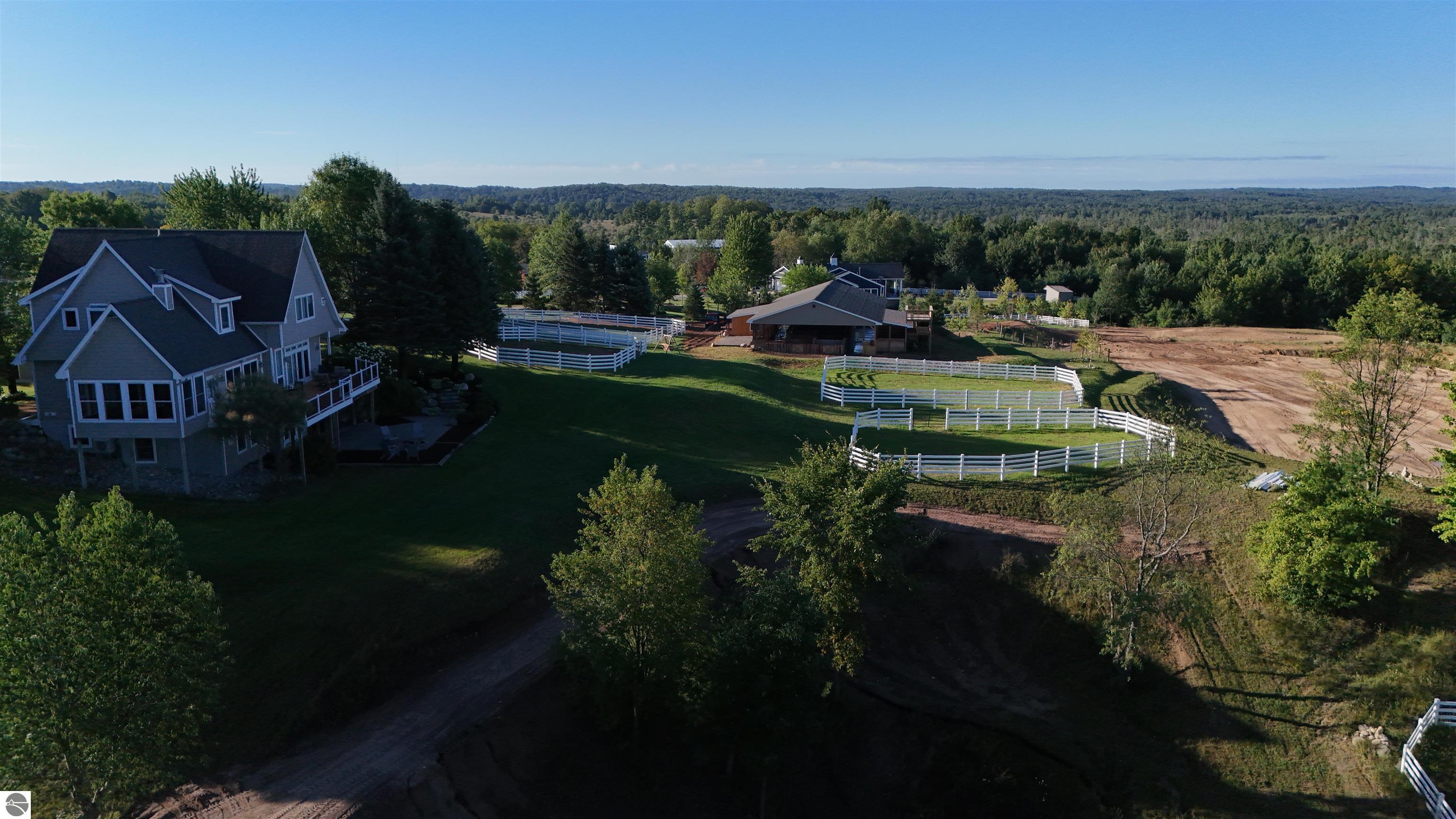 Aerial view of a gray house, stable, and white-fenced paddocks surrounded by woods.