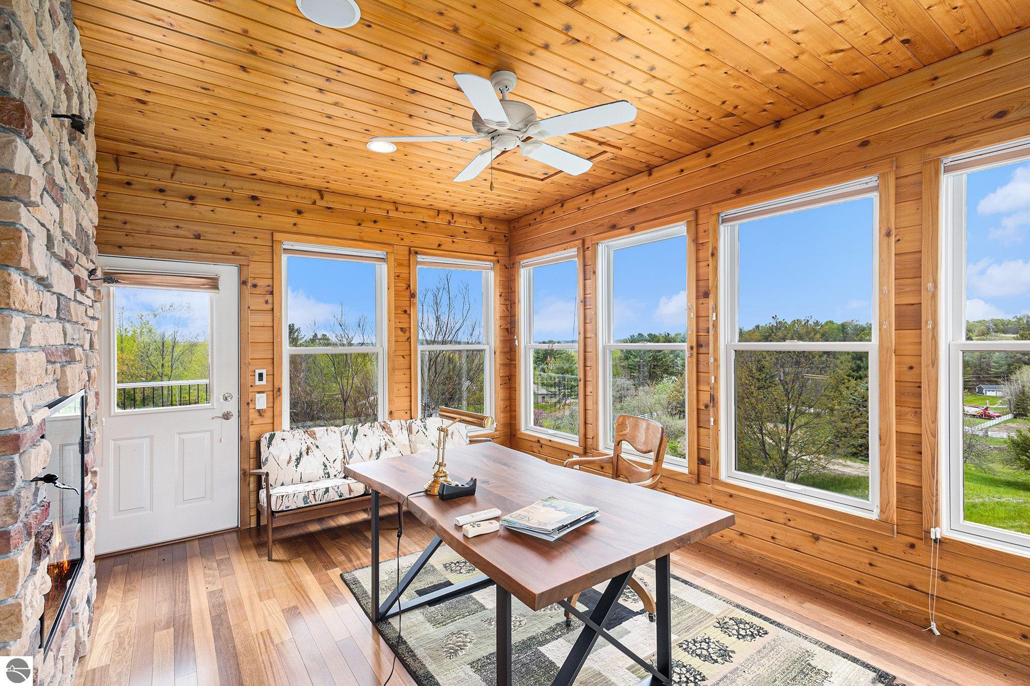 Wood-paneled sunroom with a stone fireplace, large windows, and a table overlooking trees.
