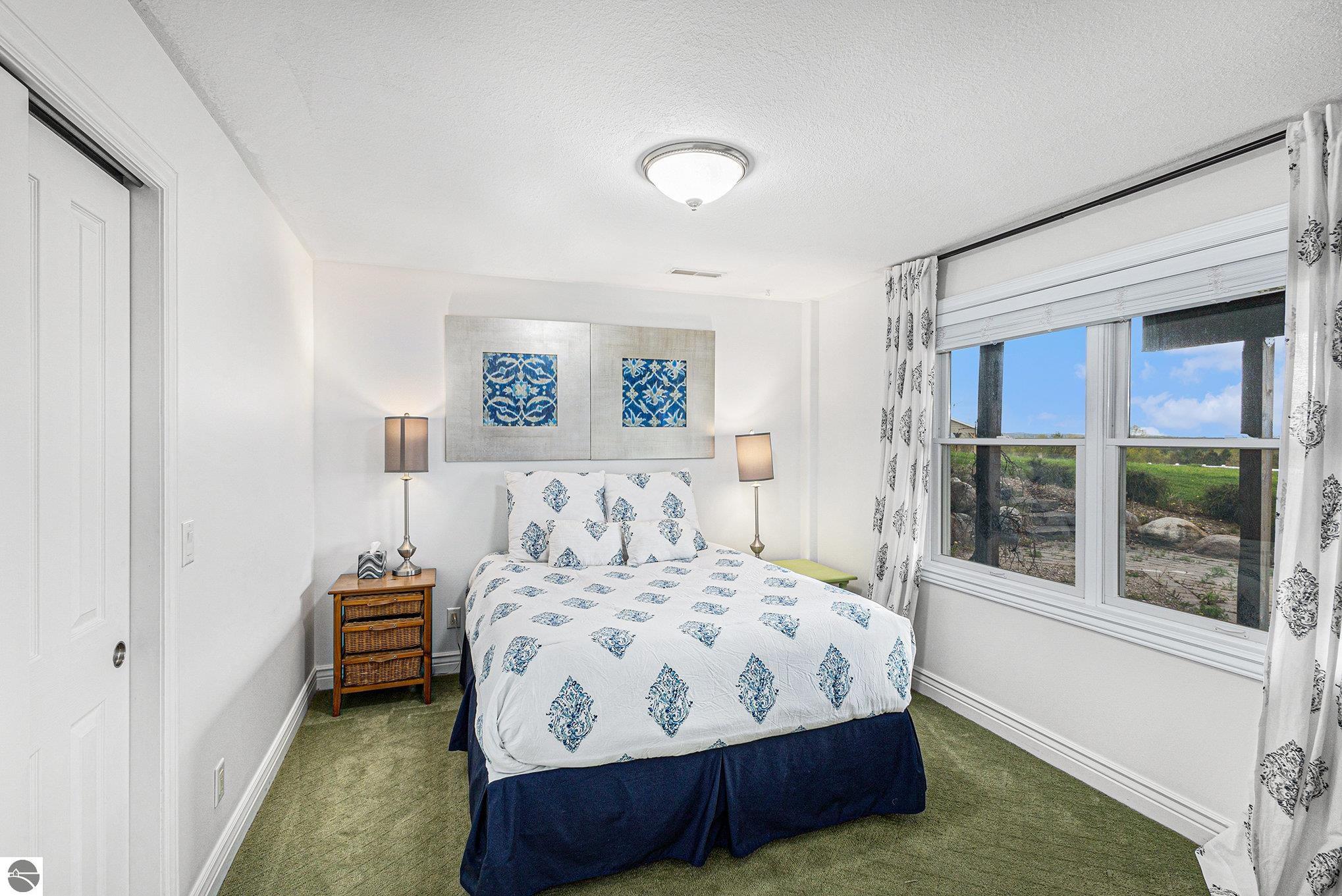 Bedroom with a patterned bed, matching bedside lamps, and a large window overlooking a grassy yard.