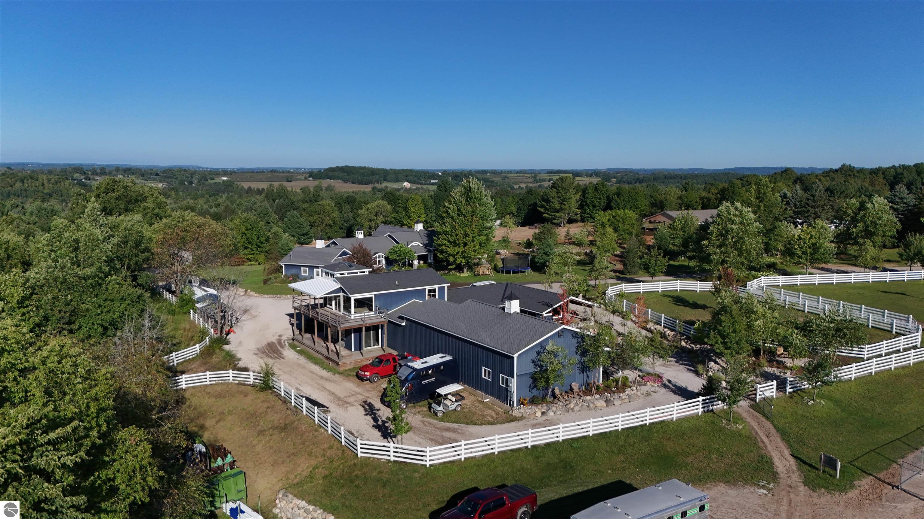 Aerial view of a blue home and equestrian property with white-fenced paddocks and wooded surroundings.