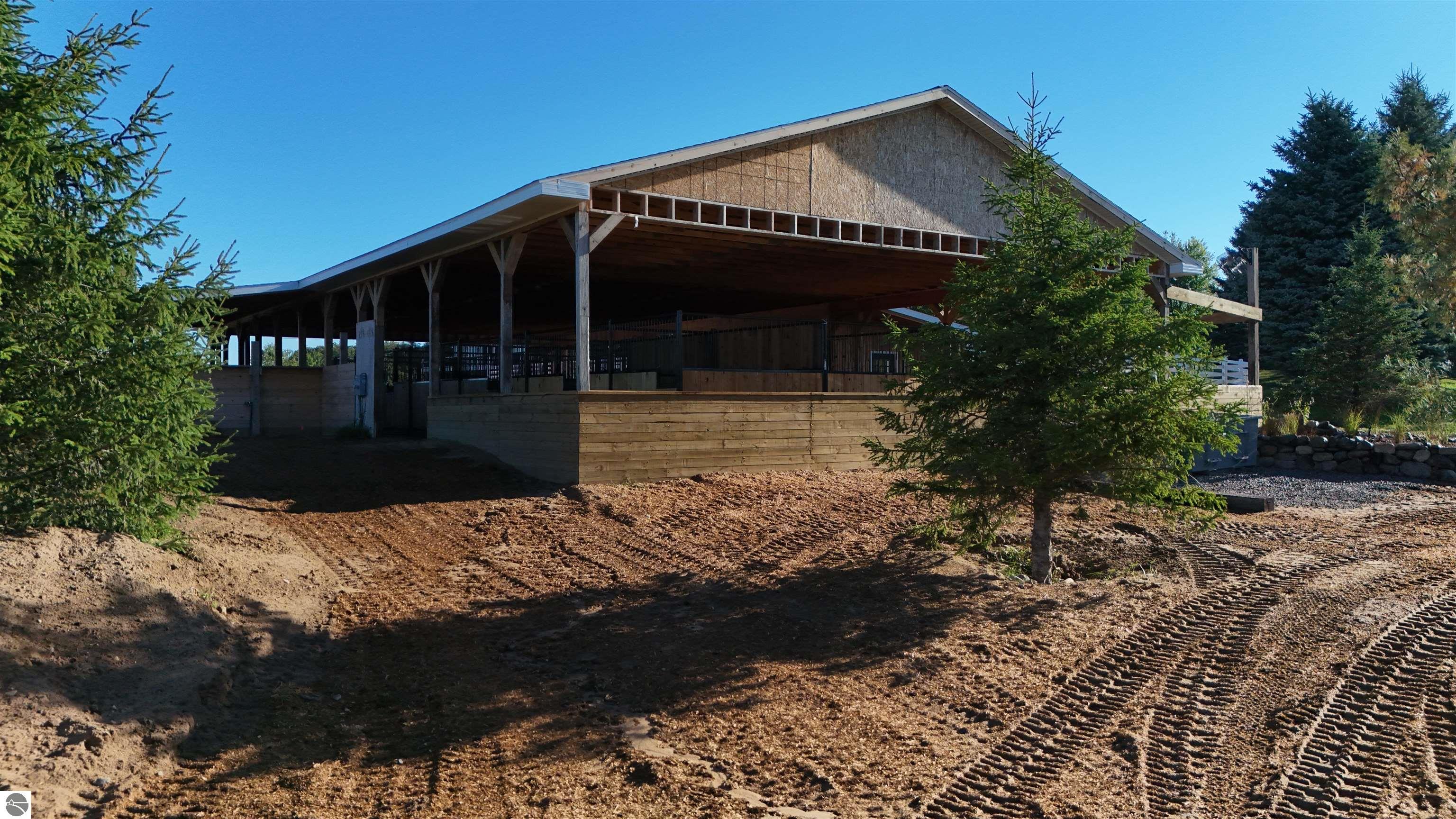 Timber-framed equestrian barn with a broad covered roof, enclosed stalls, and evergreen trees.