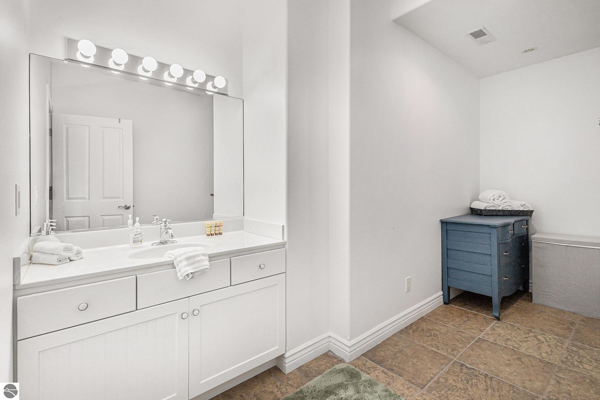 Bright bathroom with a white vanity and mirror, brown tile flooring, and a blue chest with folded towels.