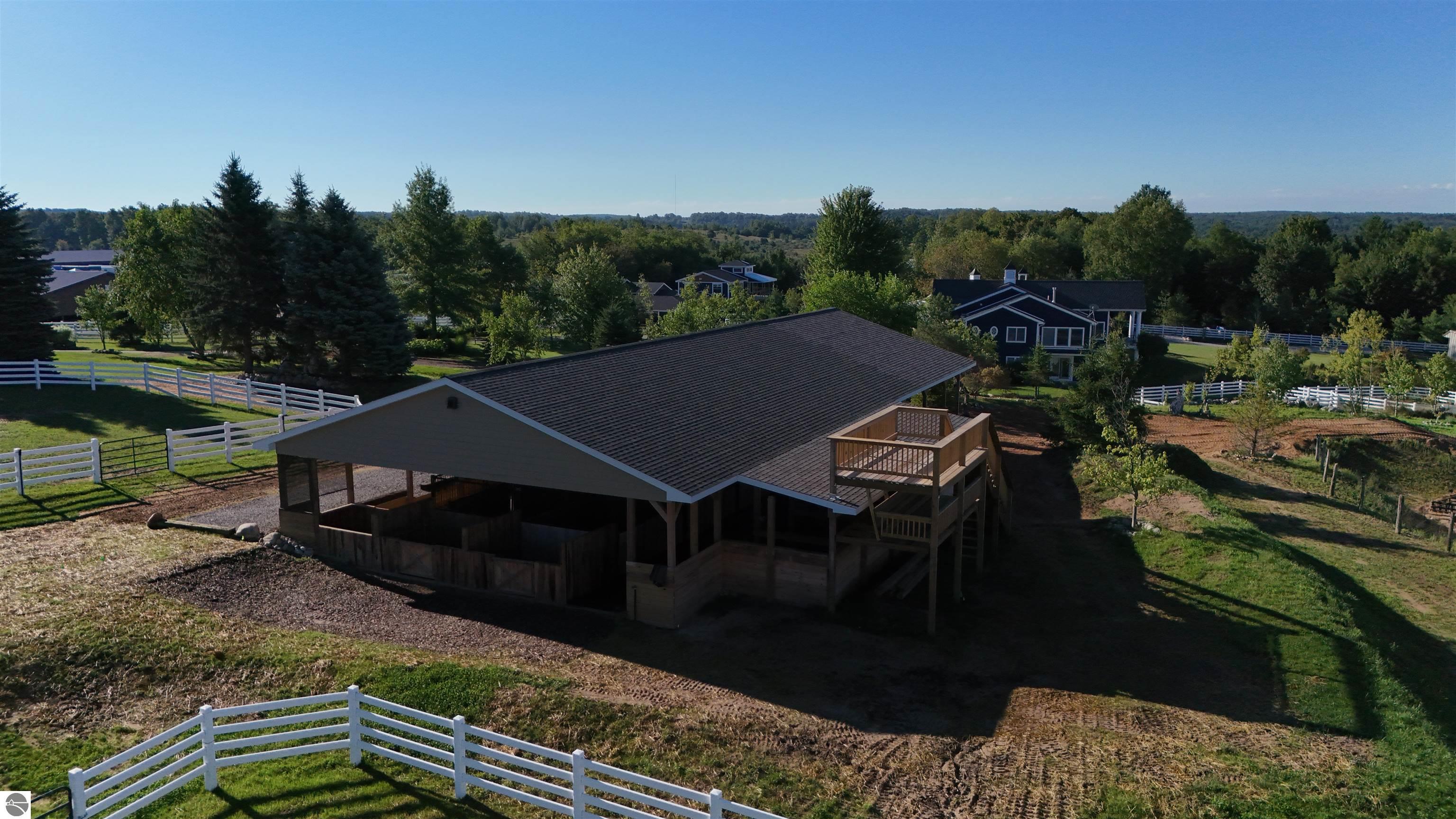 Aerial view of a large equestrian barn with white fencing and wooded grounds.