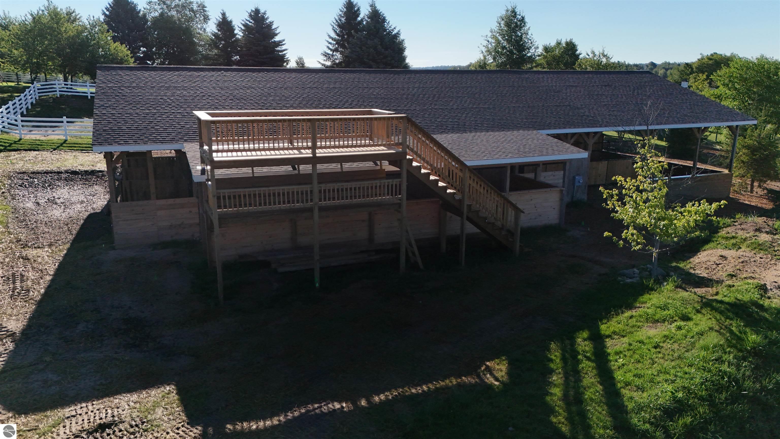 Equestrian barn with a raised wooden viewing platform and stairs, surrounded by fencing and trees.