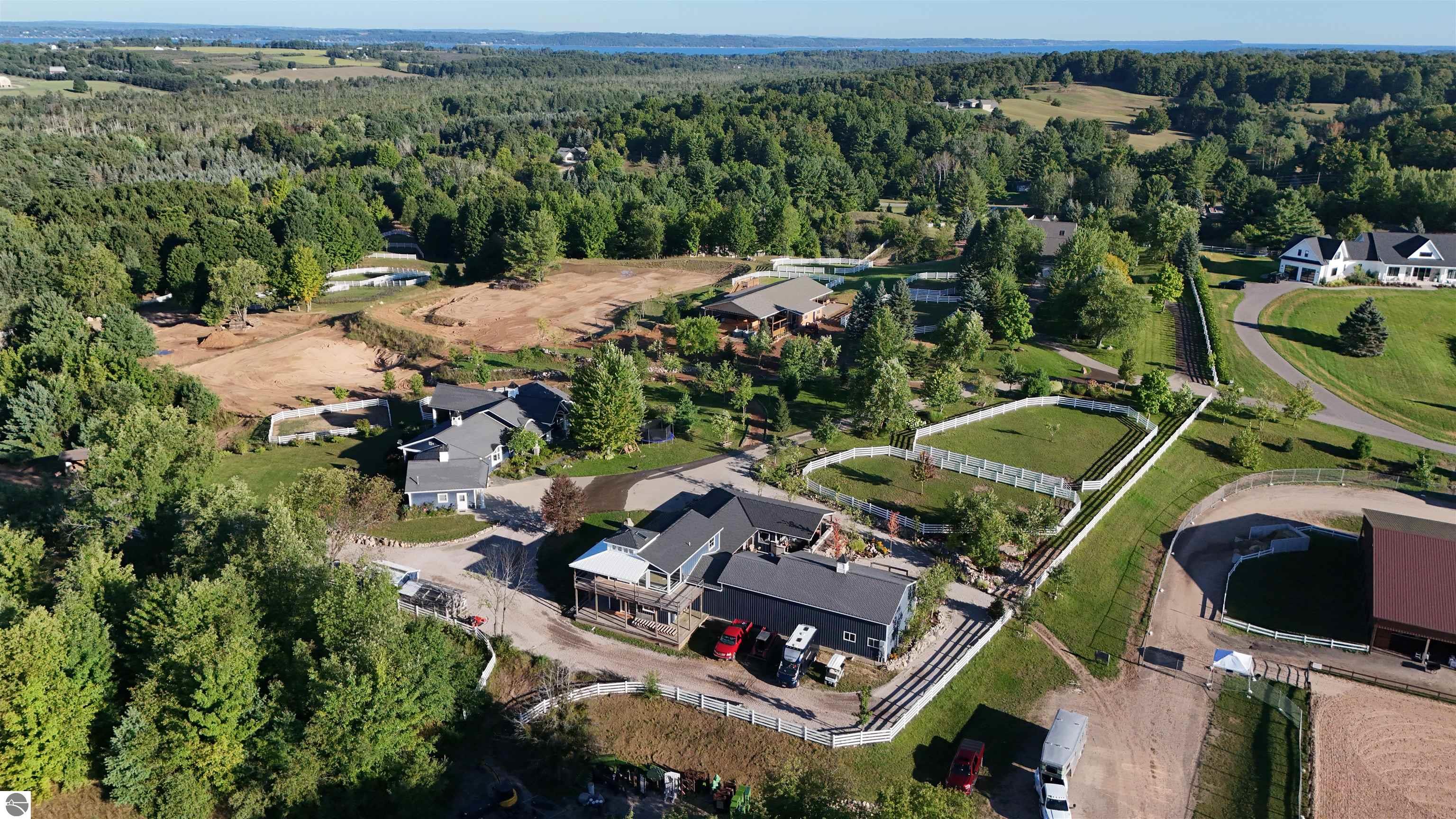 Aerial view of an equestrian estate with white-fenced paddocks, multiple buildings, and a wooded setting.