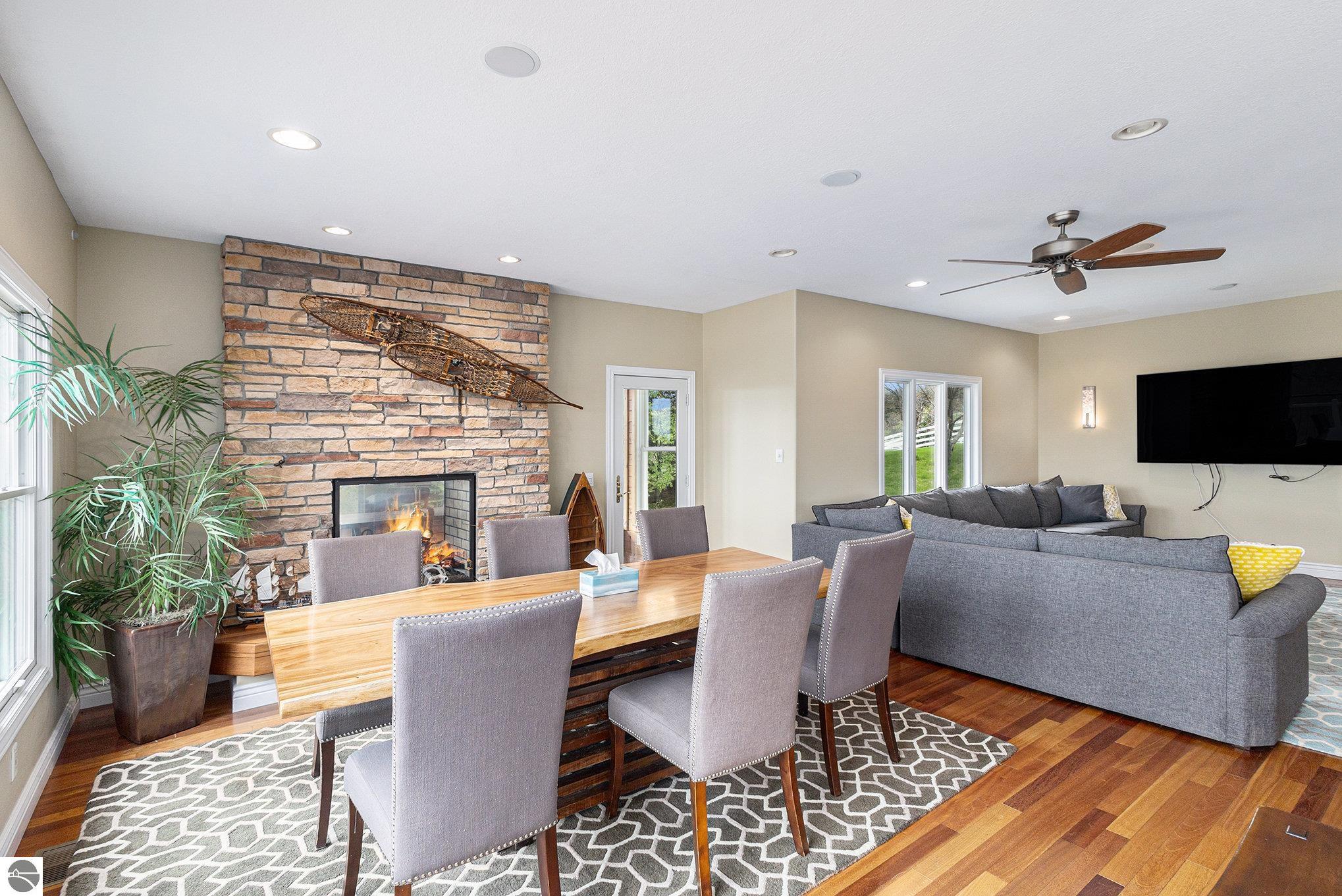 Open-plan living and dining room with a stone fireplace, wood table, gray sectional, and hardwood floors.