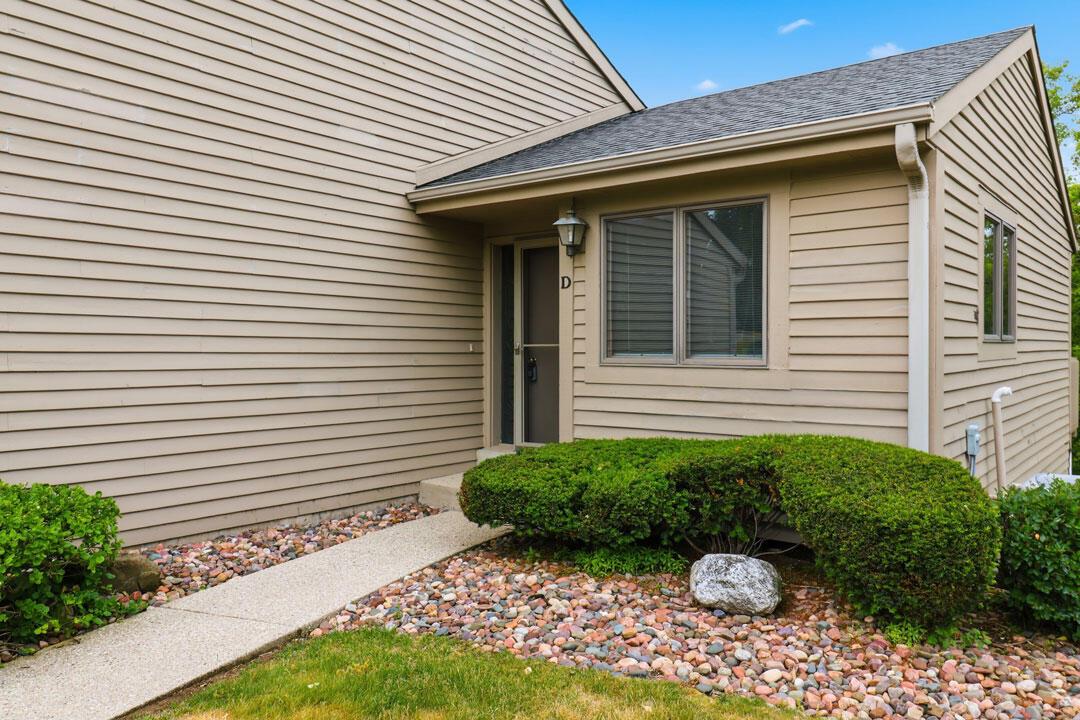 Beige-sided home entrance with a covered doorway, front window, stone landscaping, and trimmed shrubs.
