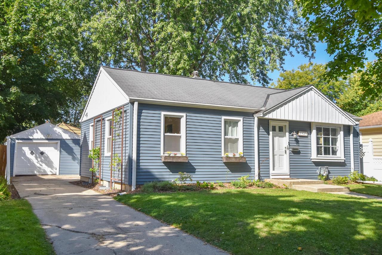 Blue single-story house with white trim, detached garage, and concrete driveway beneath mature trees.