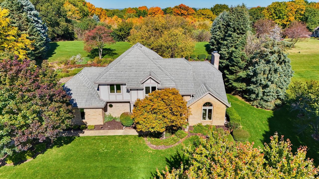 Aerial view of a brick-and-stone home with a gray roof, broad lawn, and autumn trees.