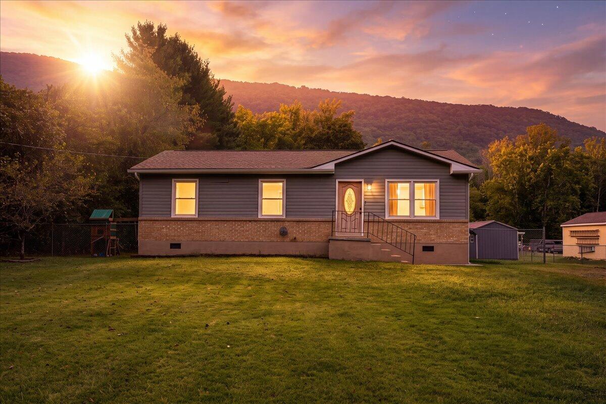 Single-story home with dark siding, brick facade, fenced lawn, and mountains behind it at sunset.