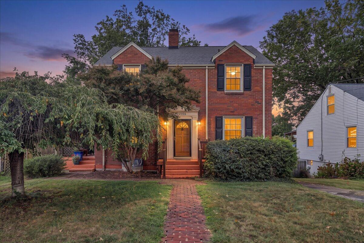 Two-story red brick house with glowing windows, dark shutters, and a brick walkway at dusk.