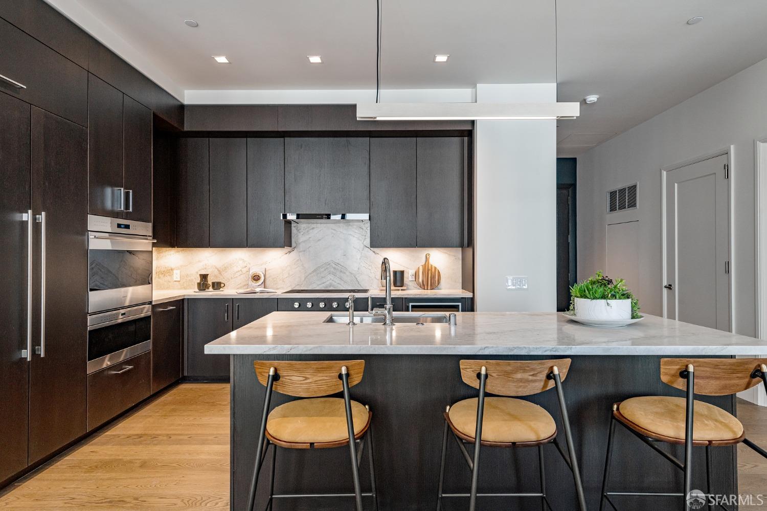 Modern kitchen with dark cabinetry, marble island and backsplash, stainless appliances, and three wooden bar stools.