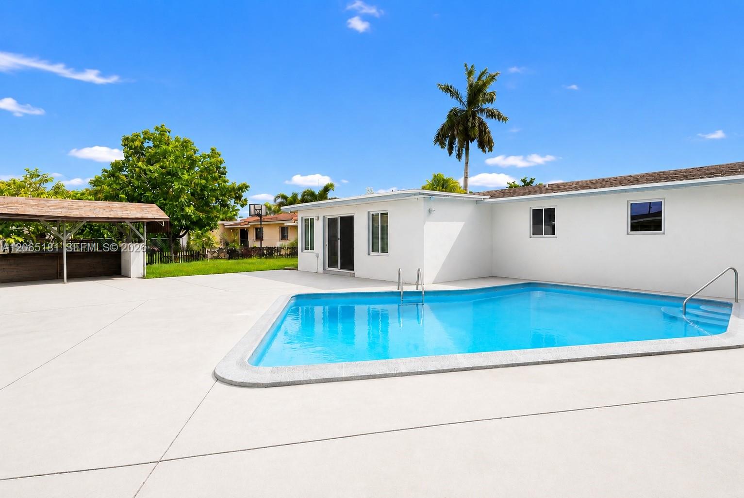 Backyard swimming pool beside a white home, with a broad concrete patio and tall palm tree.