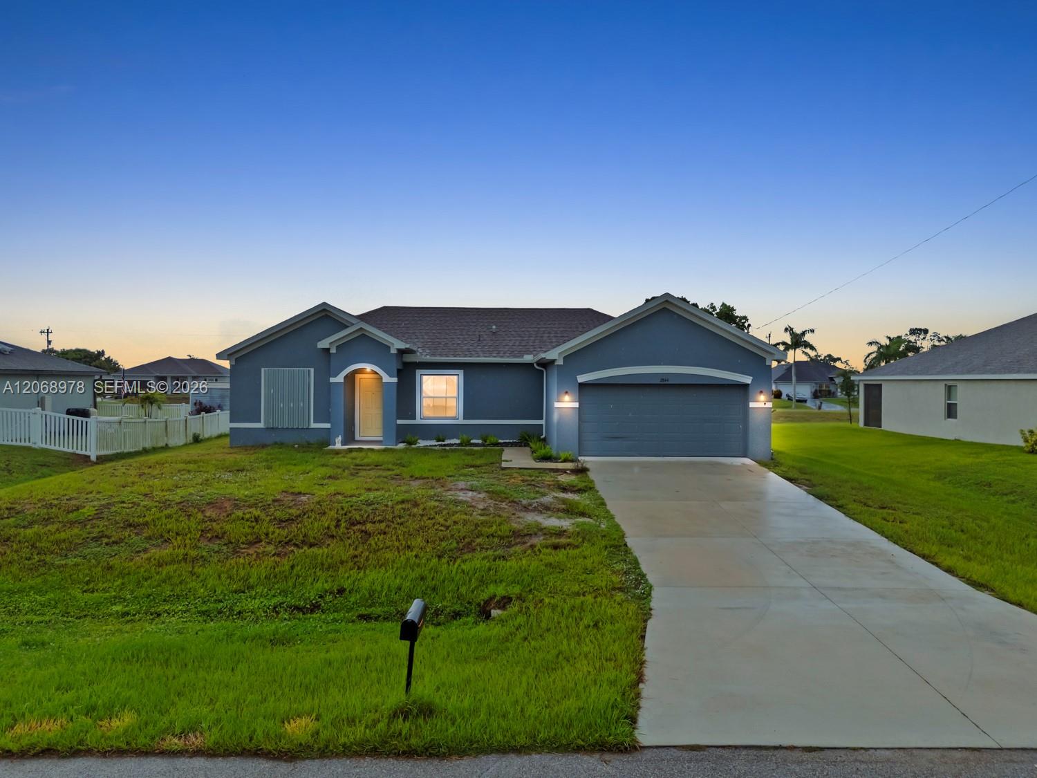 Blue single-story house with a two-car garage, broad driveway, and white fence at dusk.