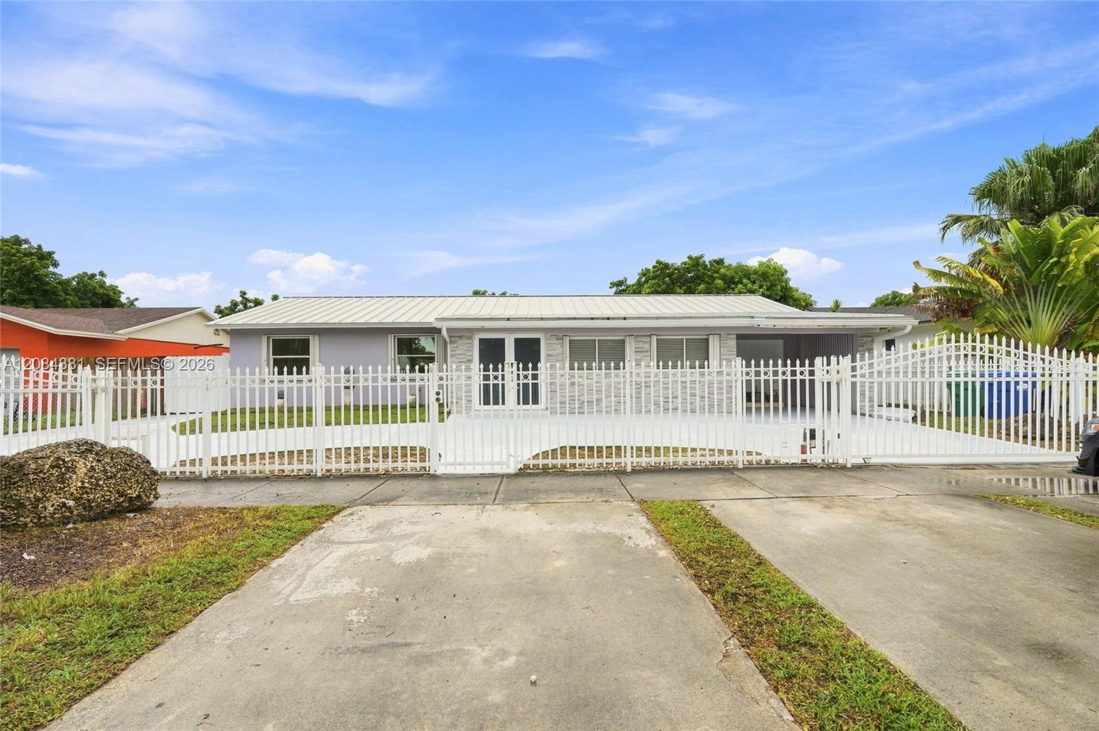 Single-story house behind a white metal fence, with a wide concrete driveway and tropical palms.