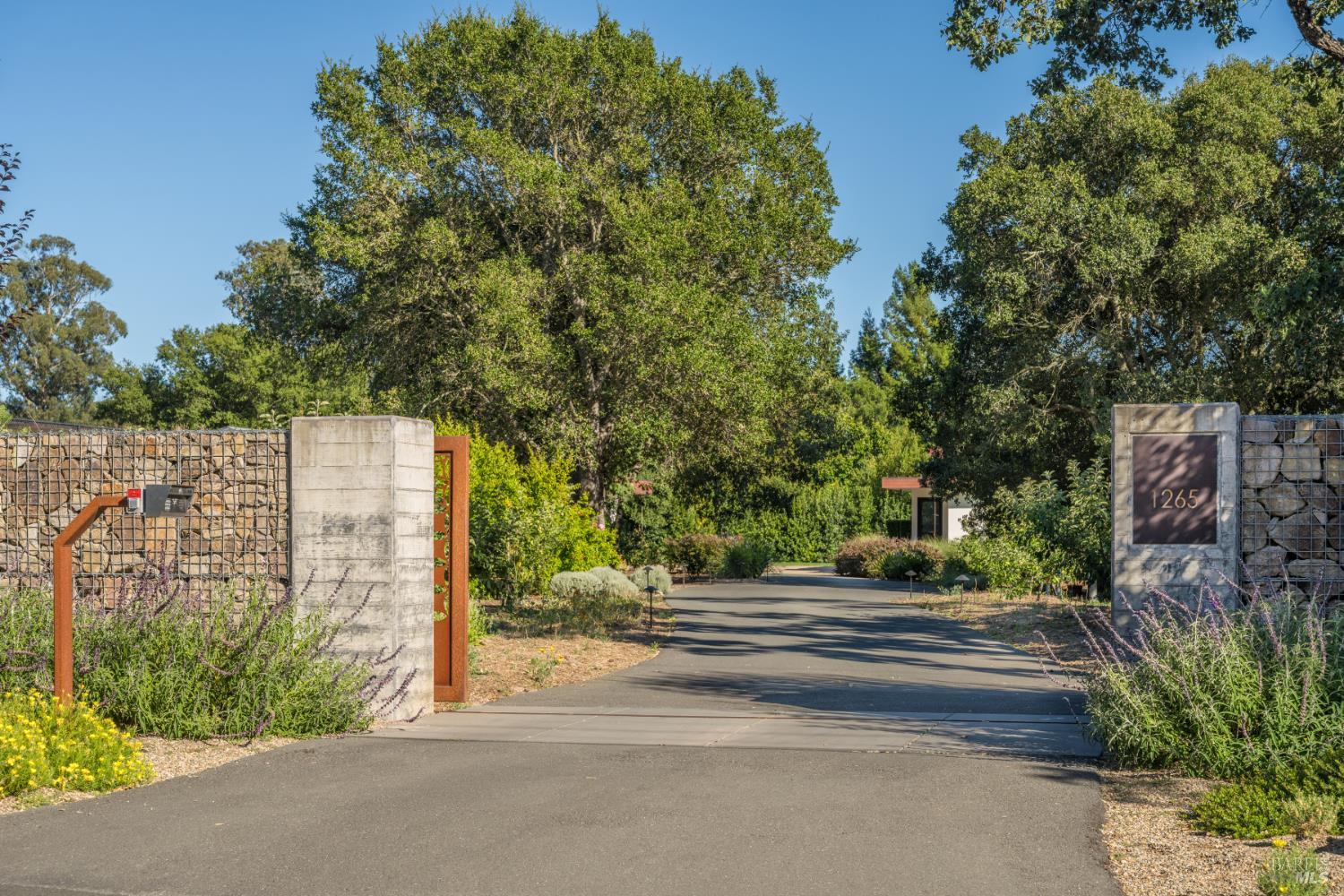 Landscaped property entrance with an open gate, gabion walls, and address 1265.