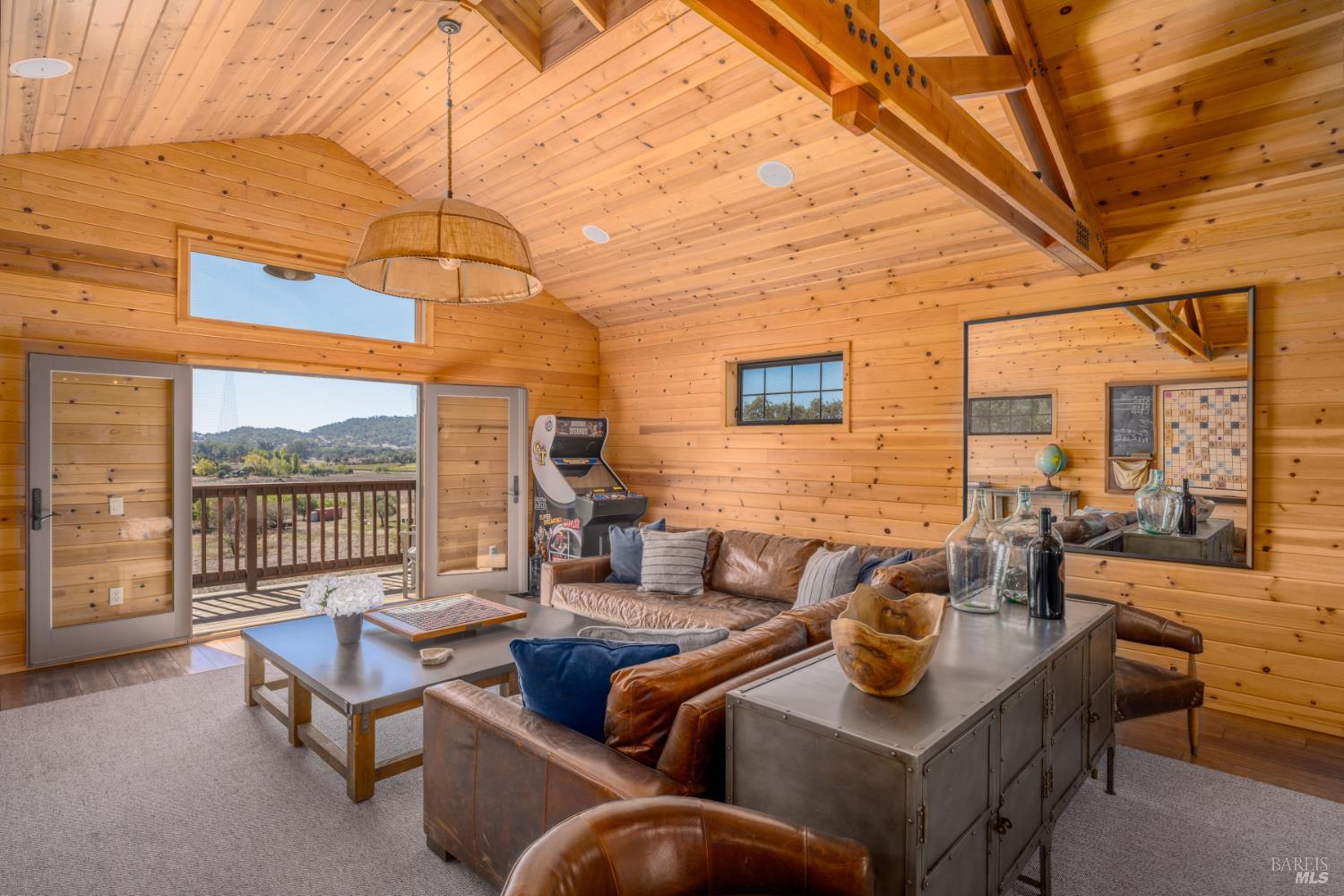 Wood-paneled living room with a vaulted timber ceiling, leather seating, arcade game, and deck access.