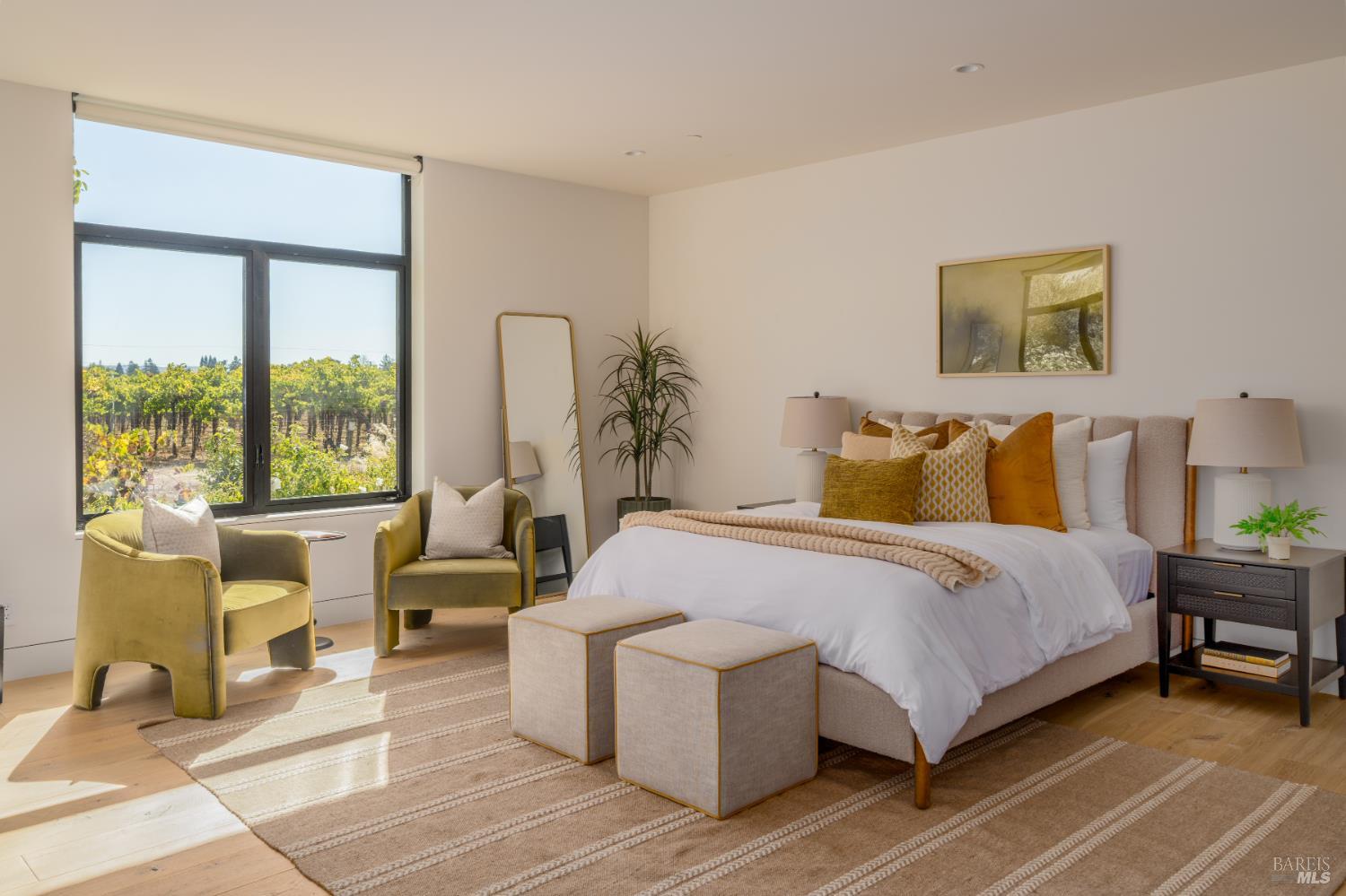 Staged bedroom with a large bed, mustard armchairs, and a window overlooking a vineyard.