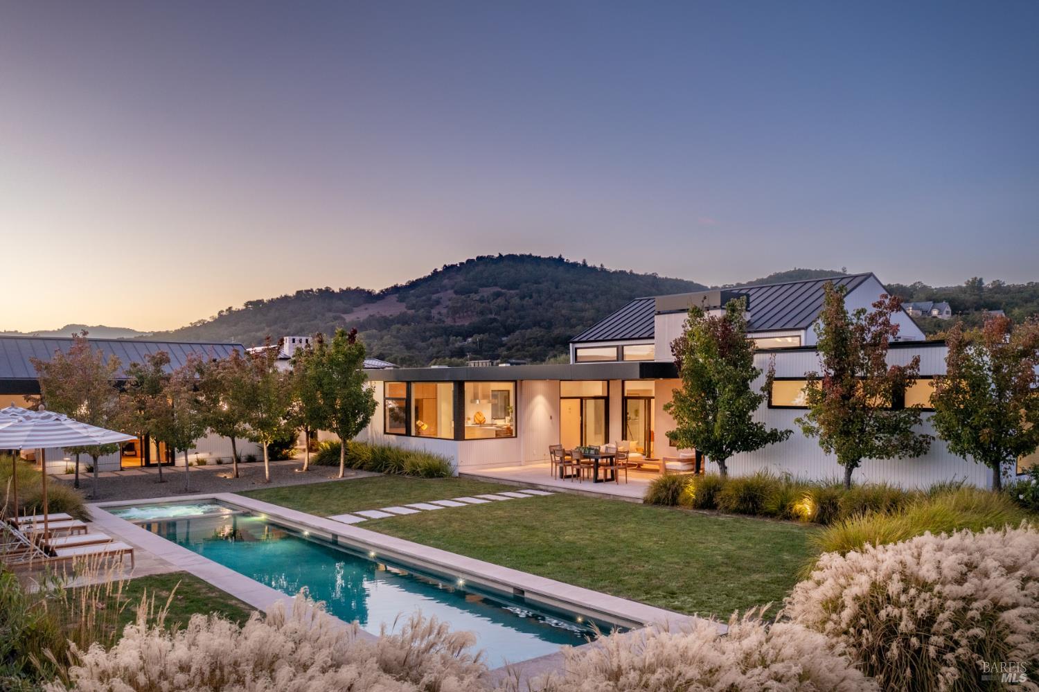 Modern white home and long pool at dusk, with wooded hills in the background.