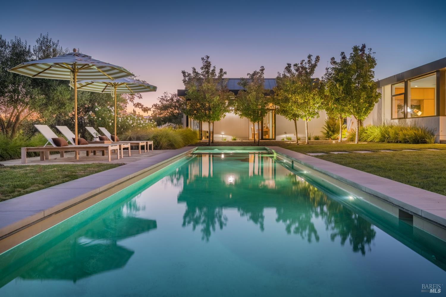 Modern home with an illuminated swimming pool, striped umbrellas, and landscaped patio at dusk.