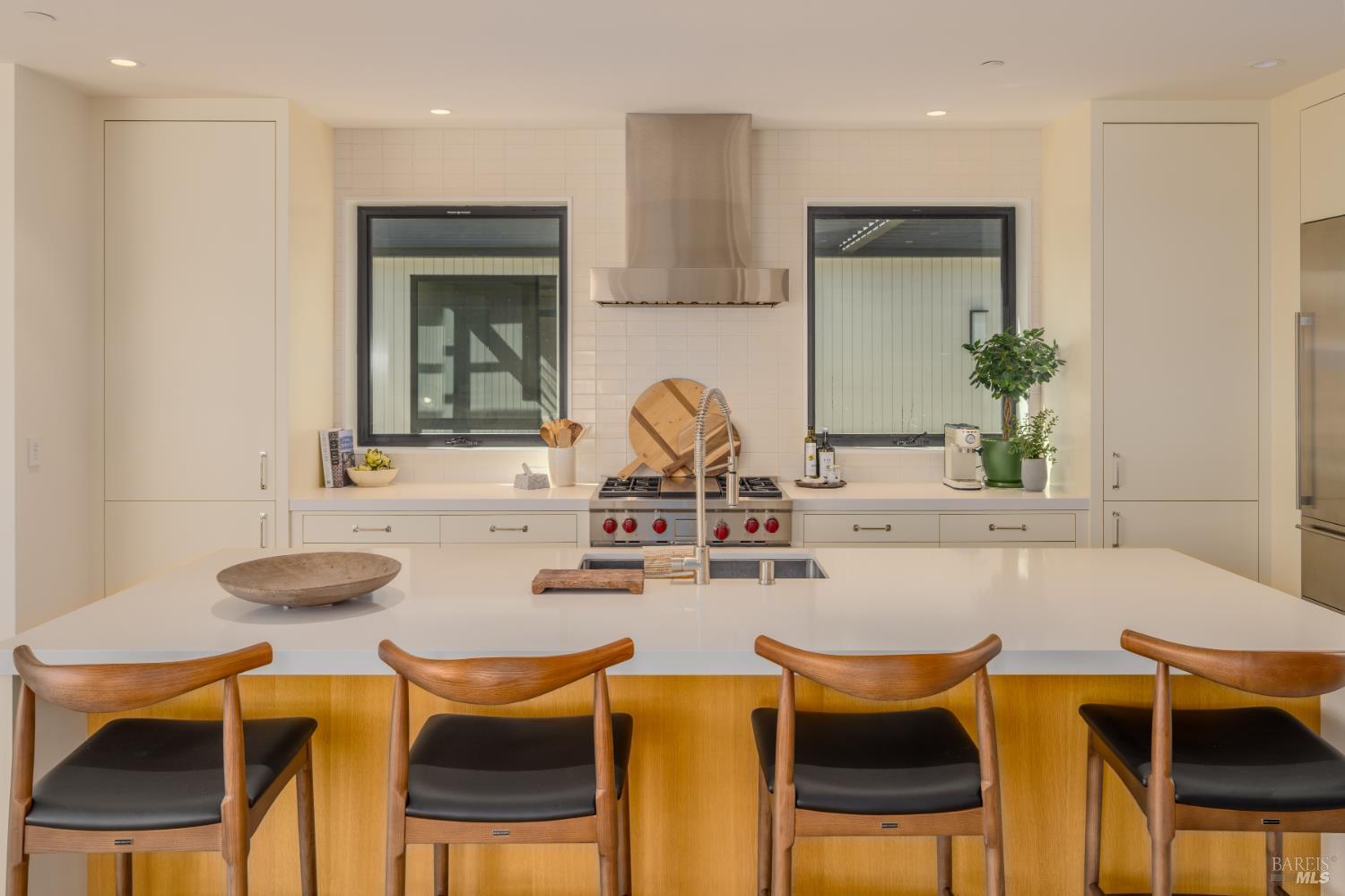 Modern kitchen with a white island, four wooden stools, and a stainless steel range beneath a broad hood.