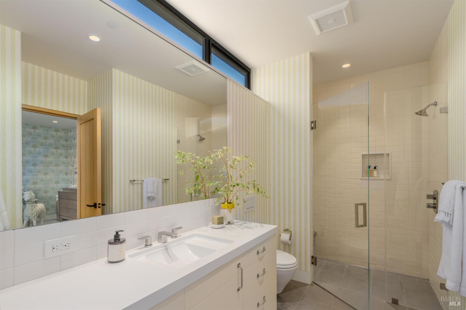 Bathroom with a long double-sink vanity, striped wallpaper, and a glass-enclosed tiled shower.