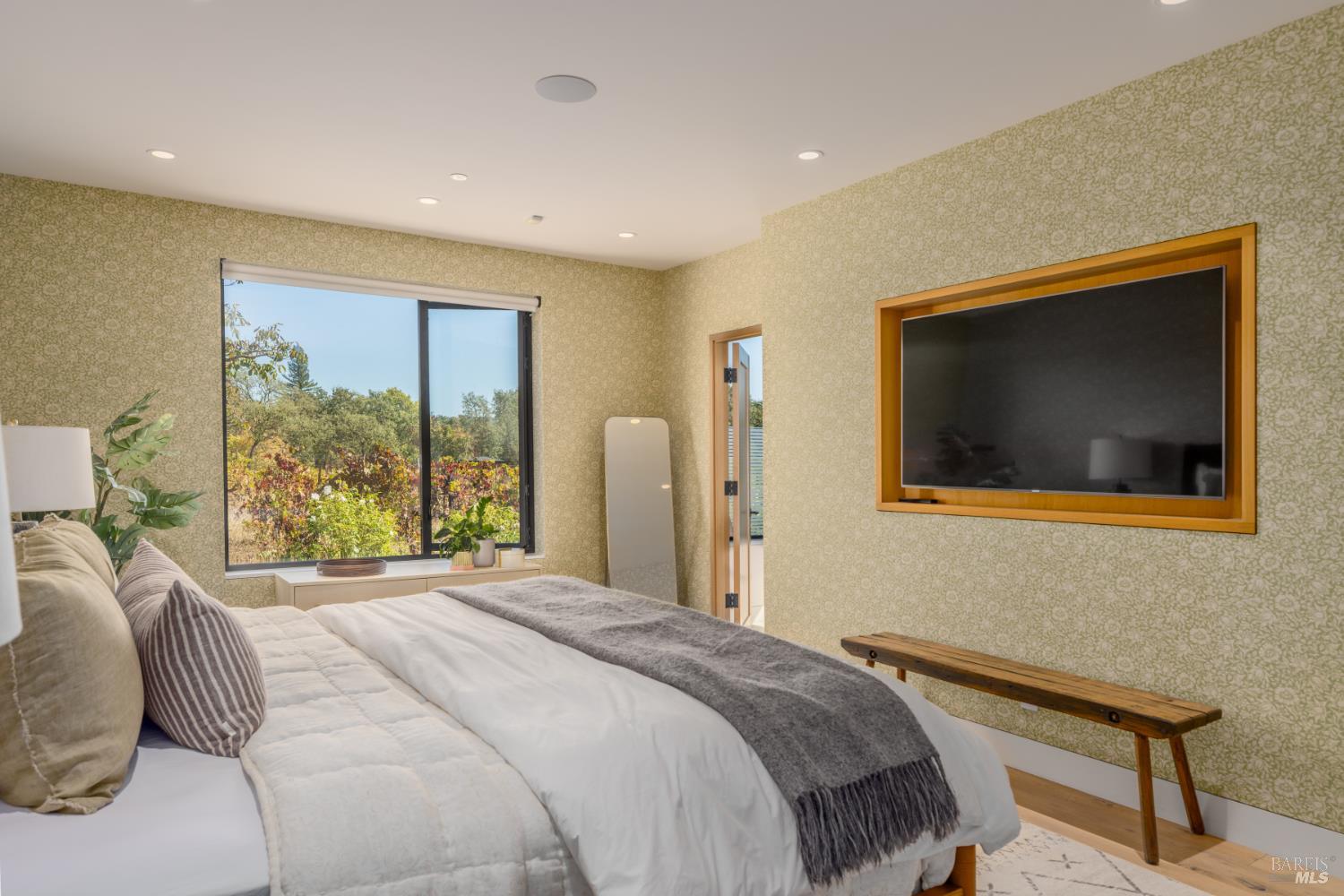 Bedroom with a large bed, patterned wallpaper, and a wide window overlooking leafy landscaping.