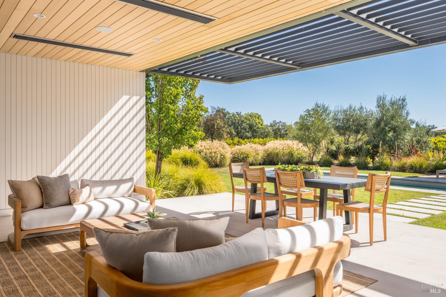 Covered patio with lounge seating and dining table beside a landscaped garden and pool.