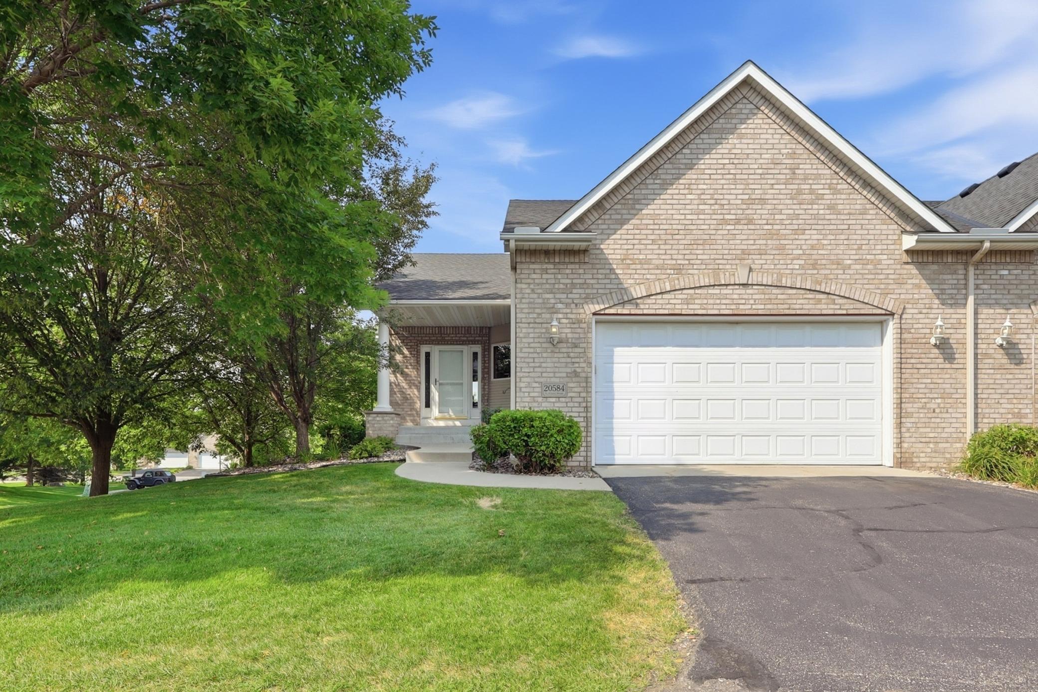 Light brick home with a gabled roof, white double garage door, covered entry, and mature trees.