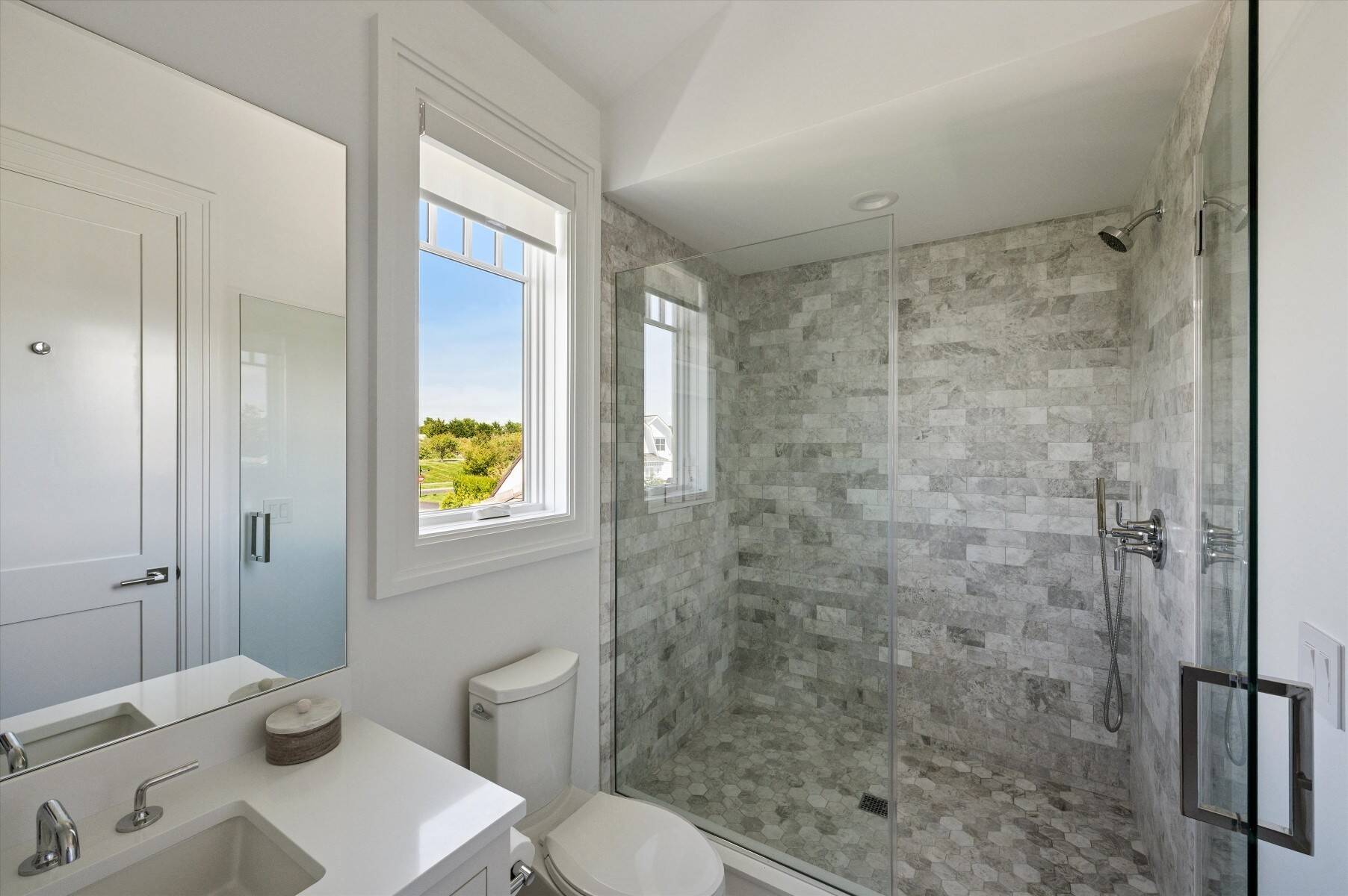 Bathroom with a glass-enclosed gray stone-tile shower, white vanity, and large window.