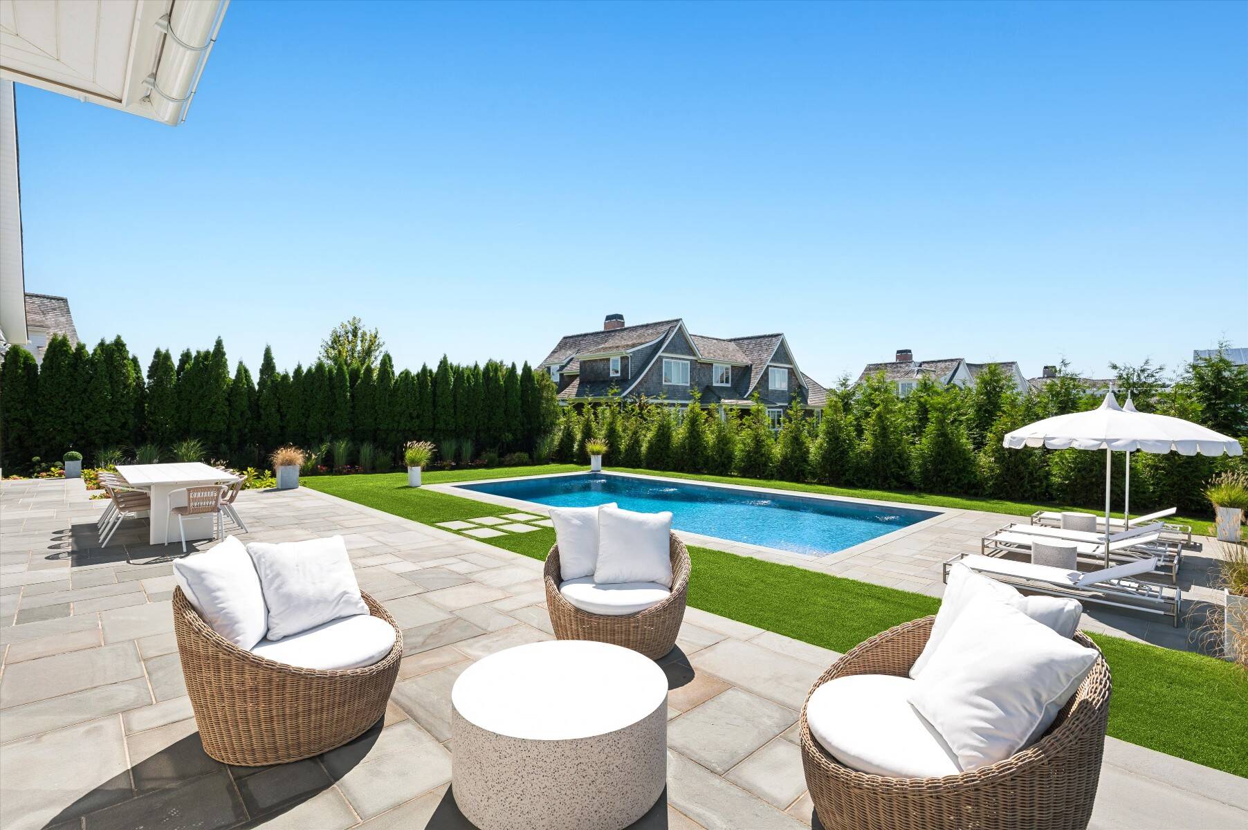 Backyard pool and stone patio with woven lounge chairs, dining table, and white umbrellas.