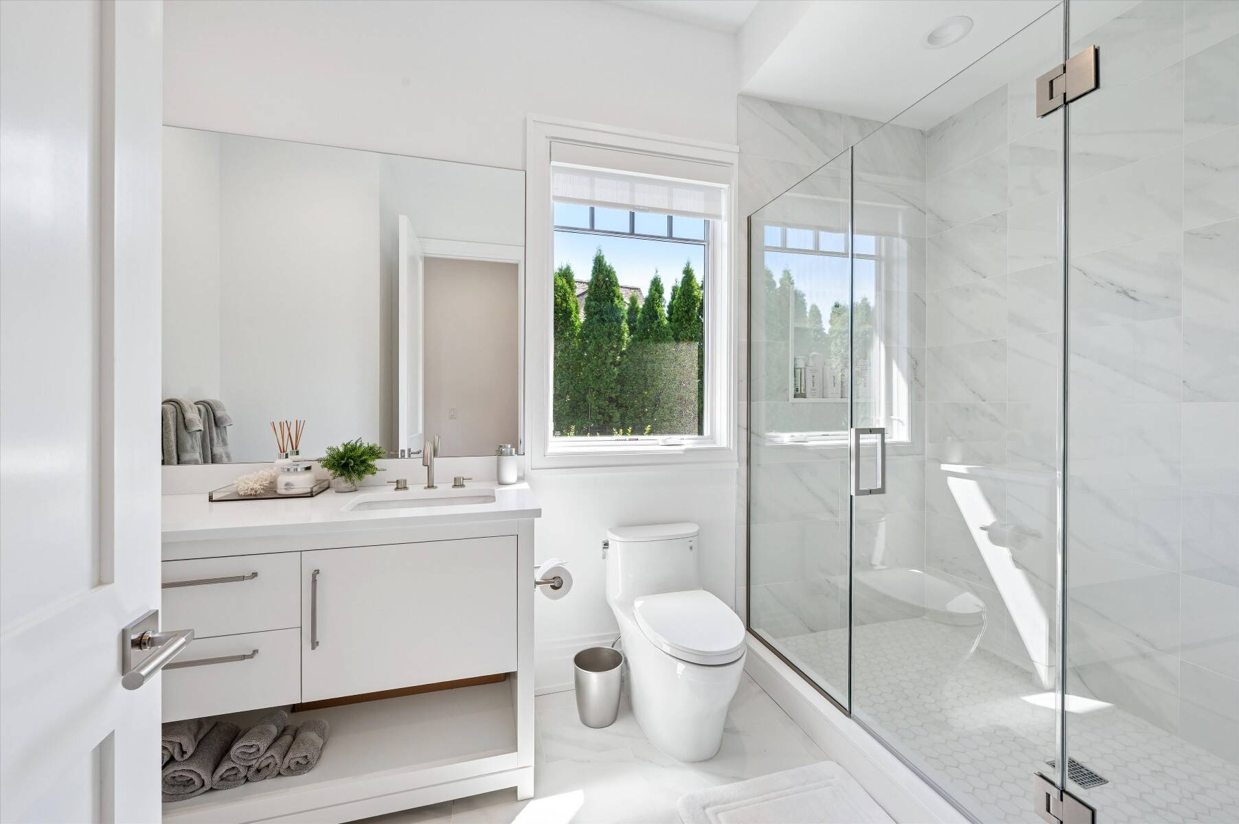 Bright bathroom with a glass-enclosed shower, white vanity, and window overlooking greenery.