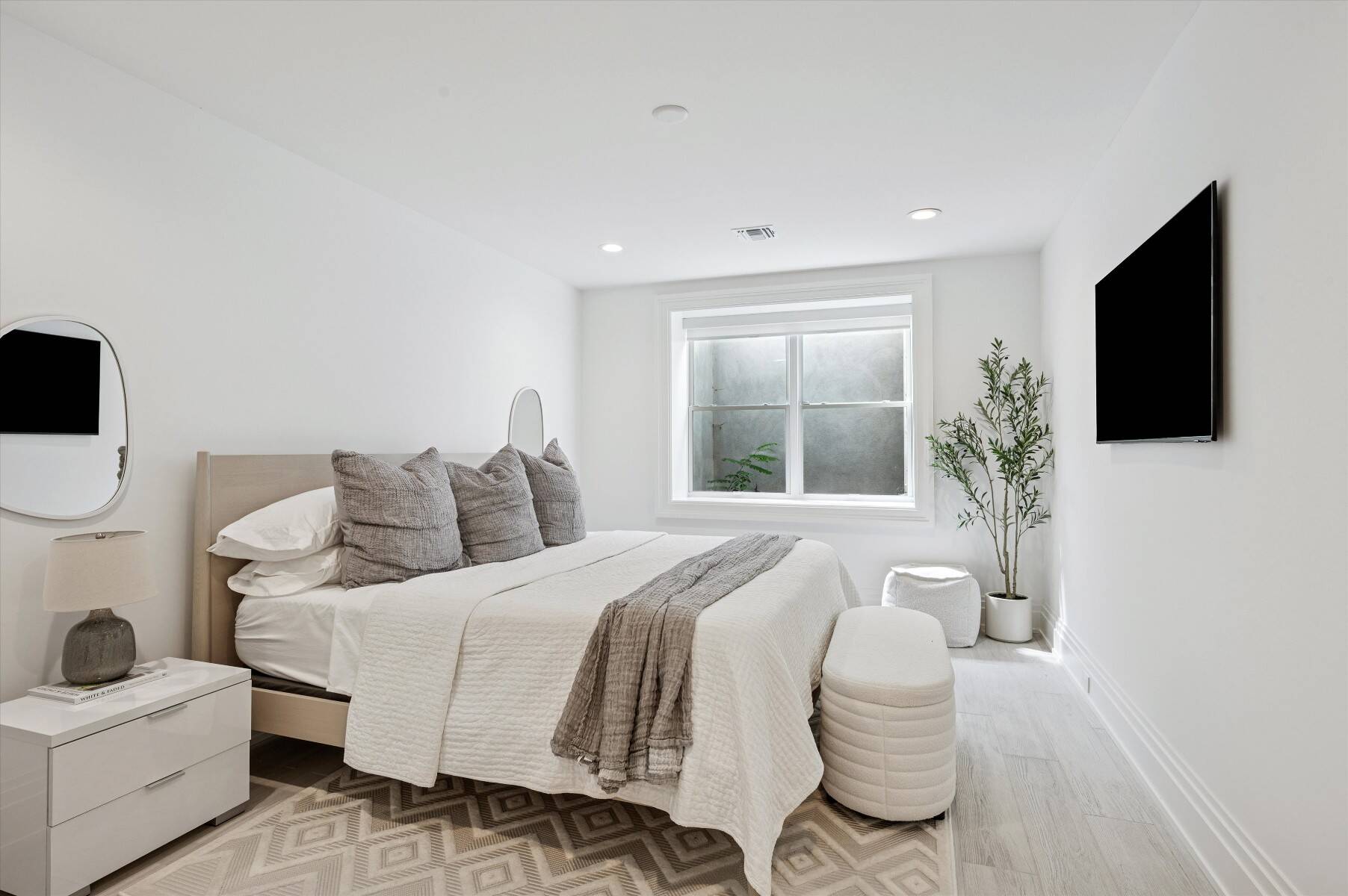 Bright neutral bedroom with a white-and-gray bed, geometric rug, and potted tree by the window.