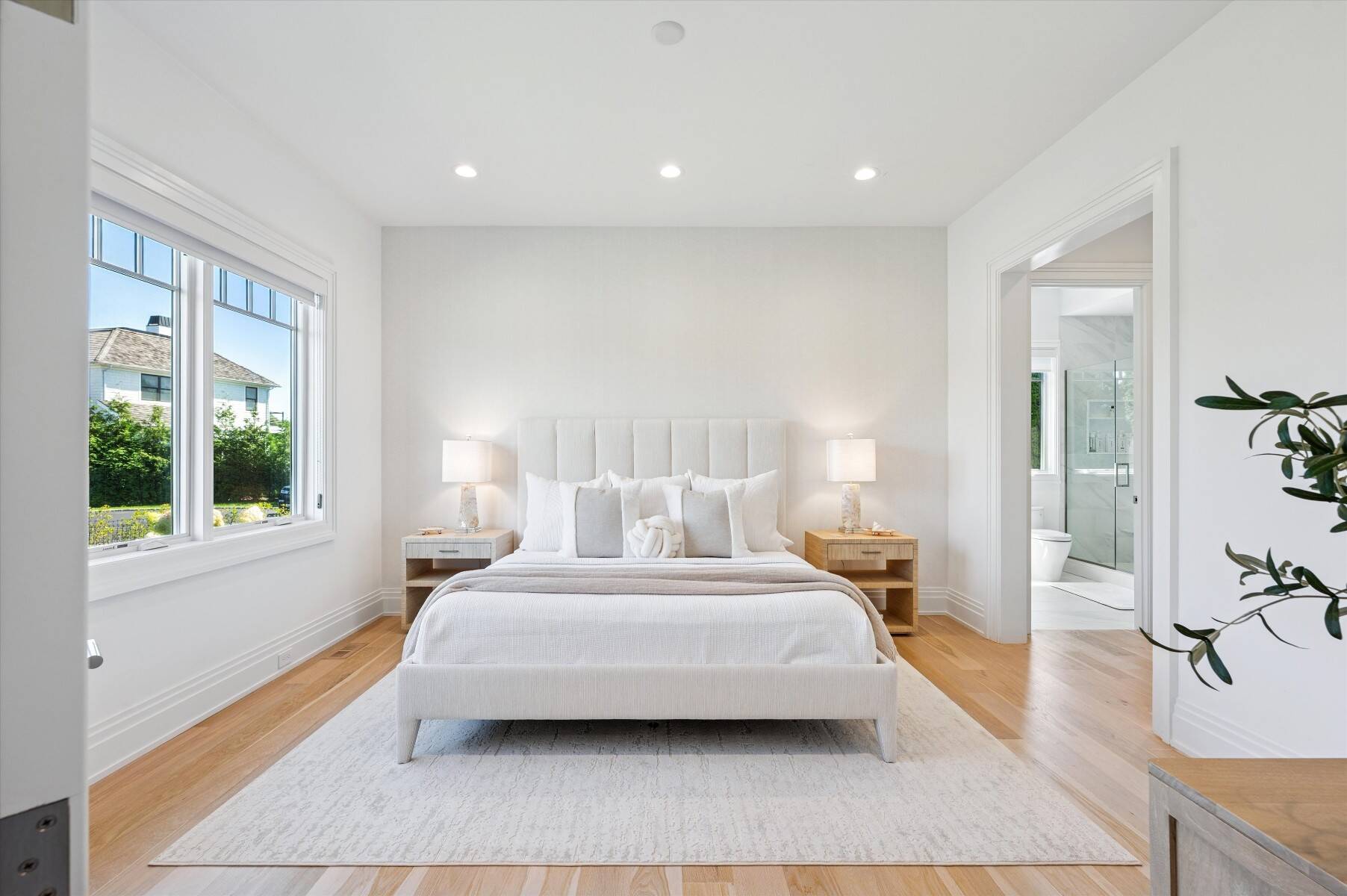 Bright bedroom with a white bed, light wood floors, large windows, and an en-suite bathroom doorway.