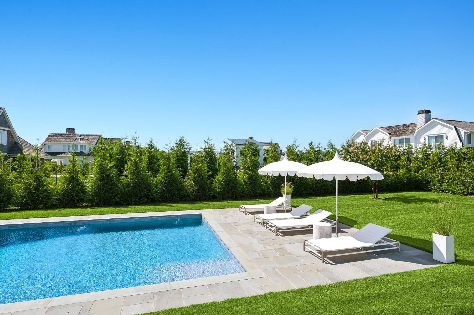 Backyard swimming pool with a stone patio, white loungers, umbrellas, and tall hedges.