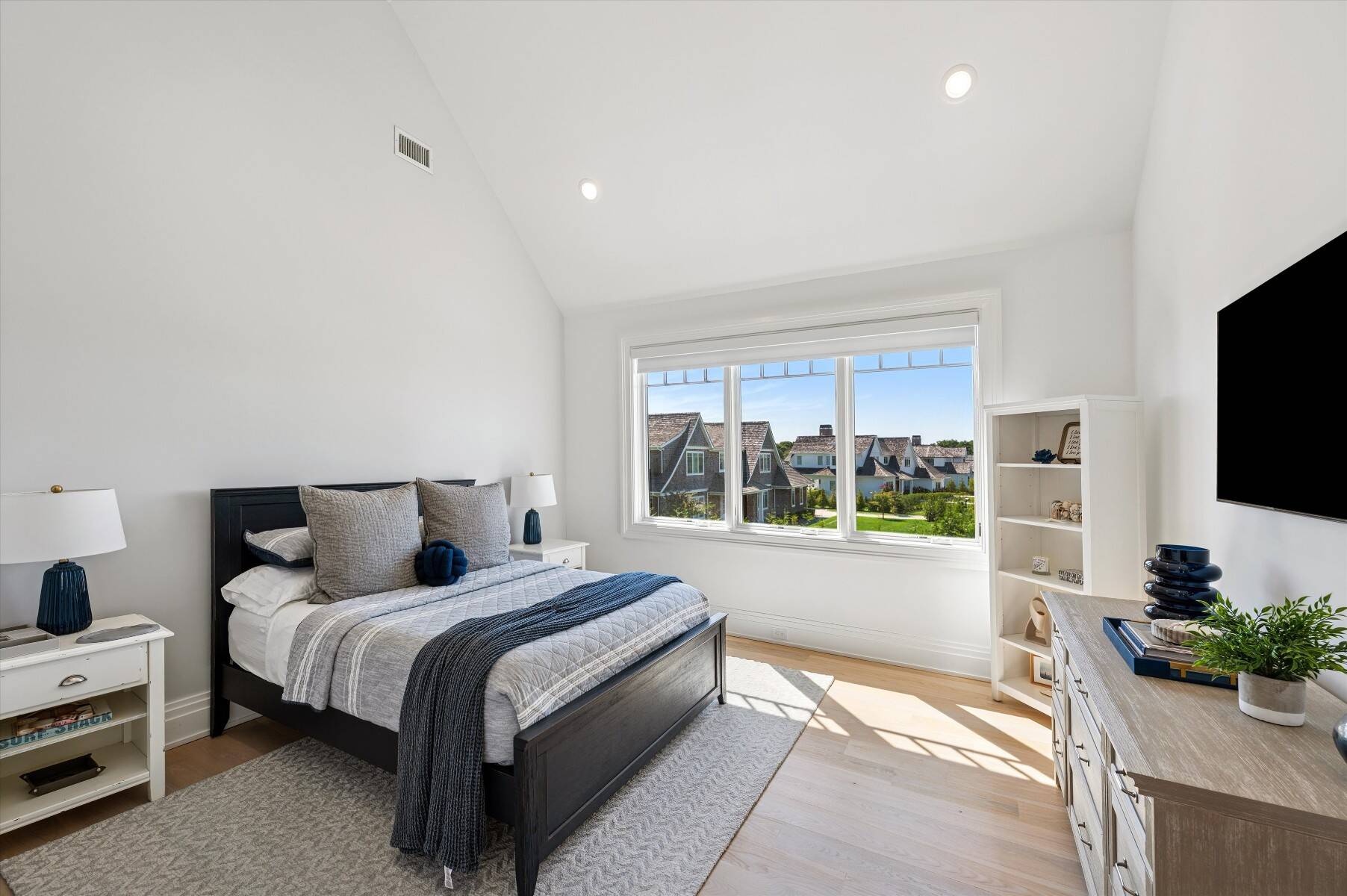 Furnished bedroom with gray and navy bedding and a large window overlooking neighboring homes.