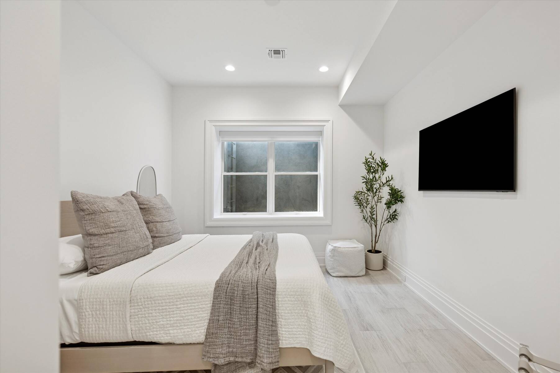 Staged bedroom with a white bed, centered window, wall-mounted television, and potted tree.