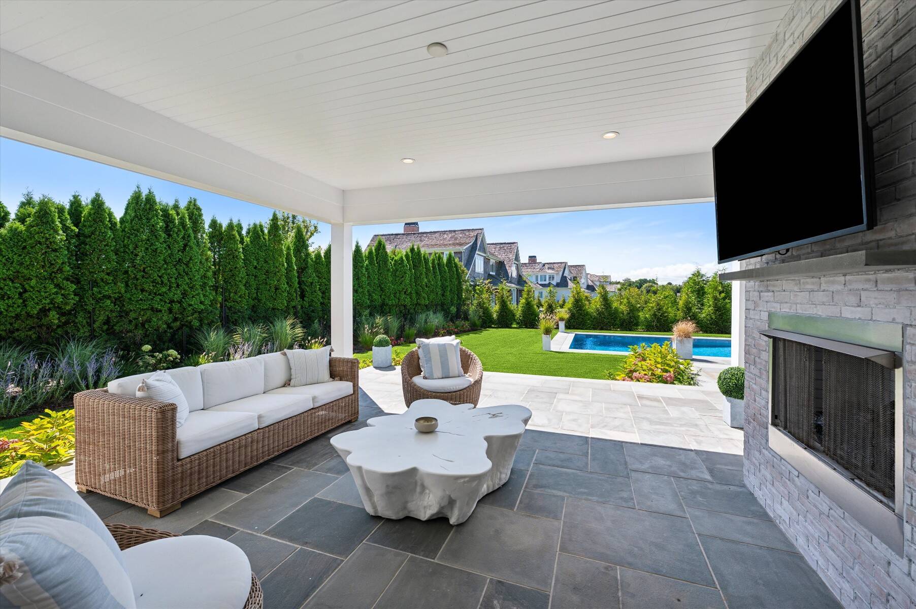 Covered patio with wicker seating and stone fireplace overlooking a lawn and swimming pool.
